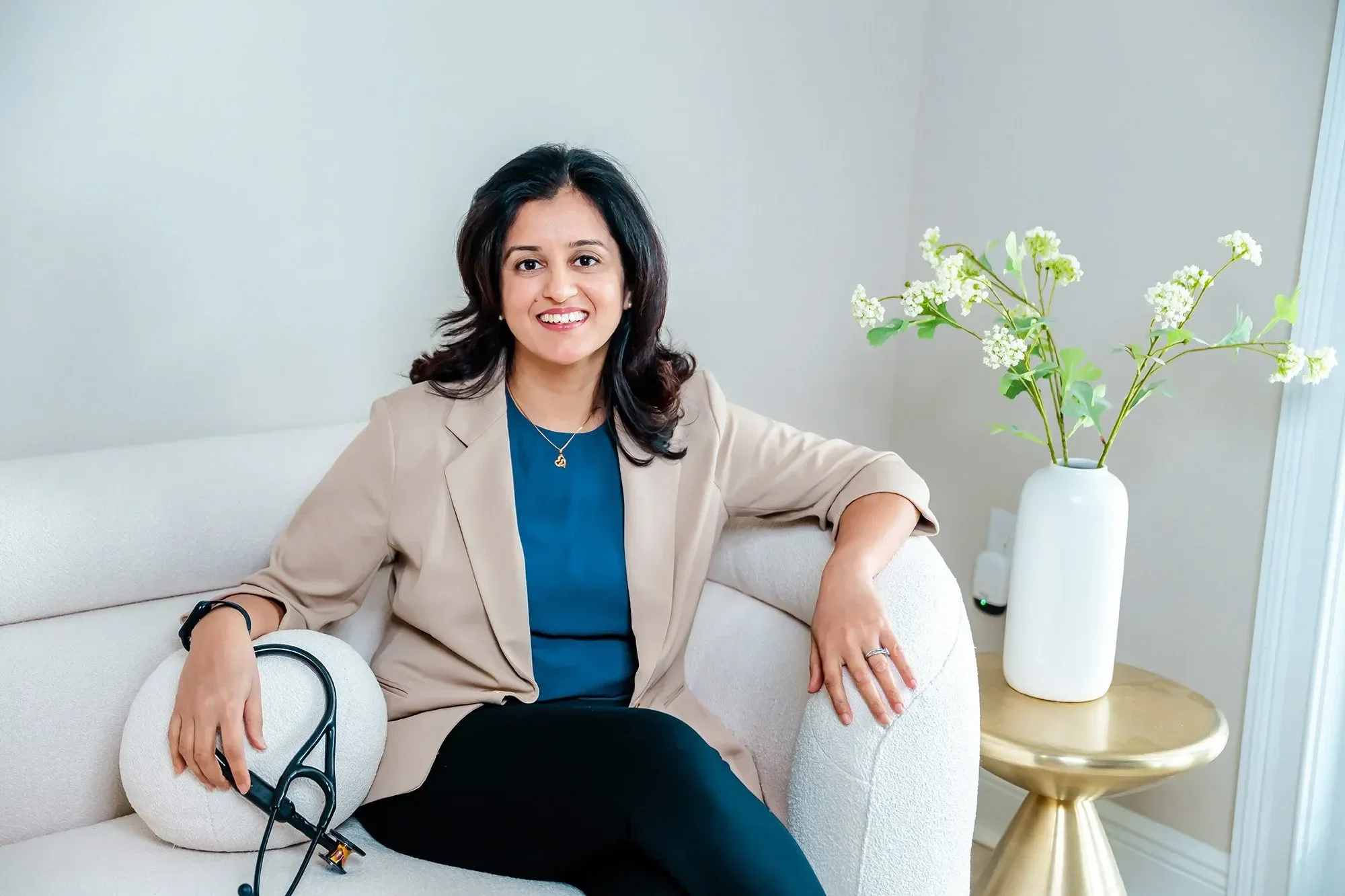 Dr. Rachita Bansal sitting on a white sofa holds a stethoscope and a heart-shaped photo frame, smiling at the camera. A vase with white flowers is on a small gold table beside her.