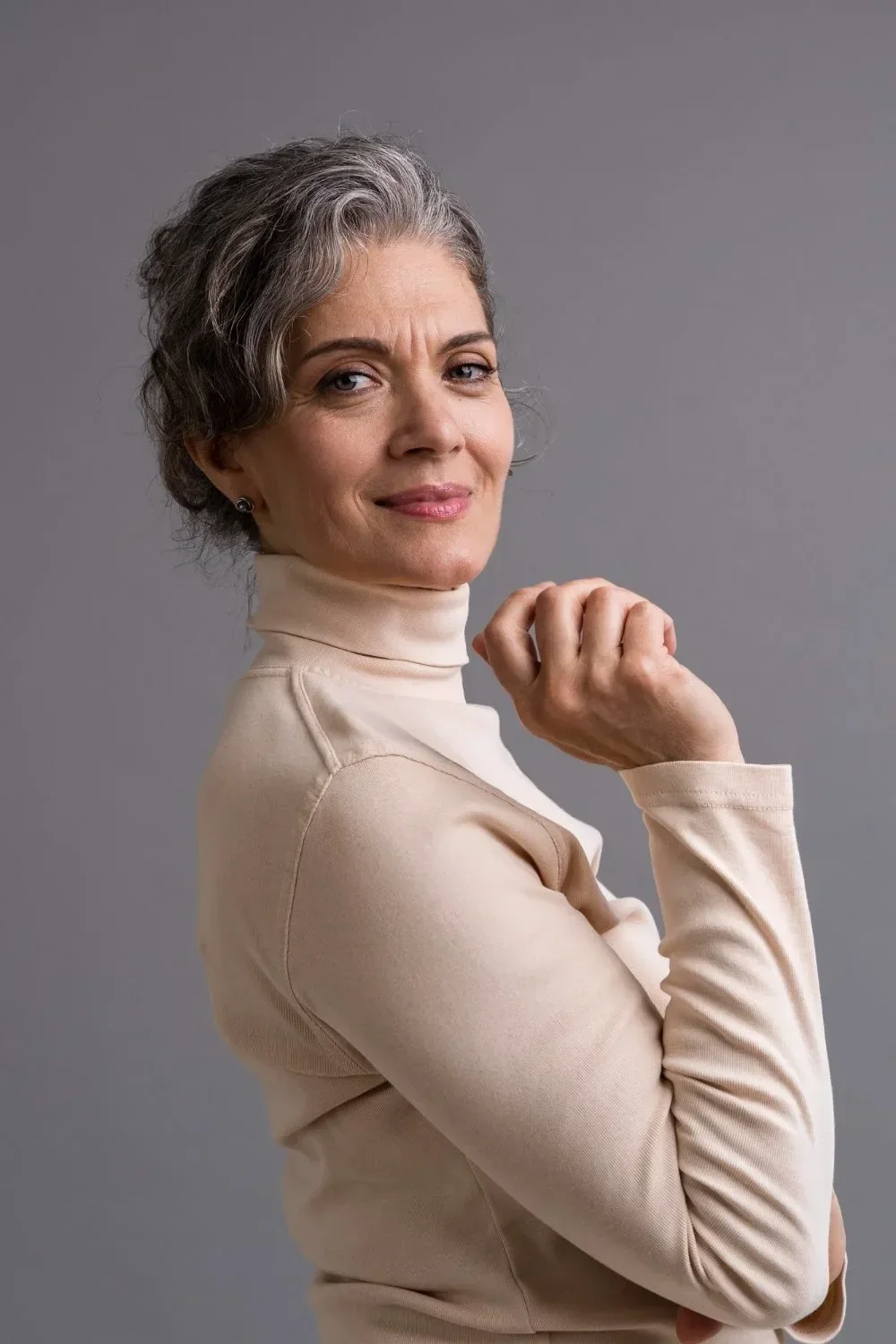 A confident middle-aged woman with short gray hair, wearing a beige turtleneck, posing with a slight smile against a plain gray background.