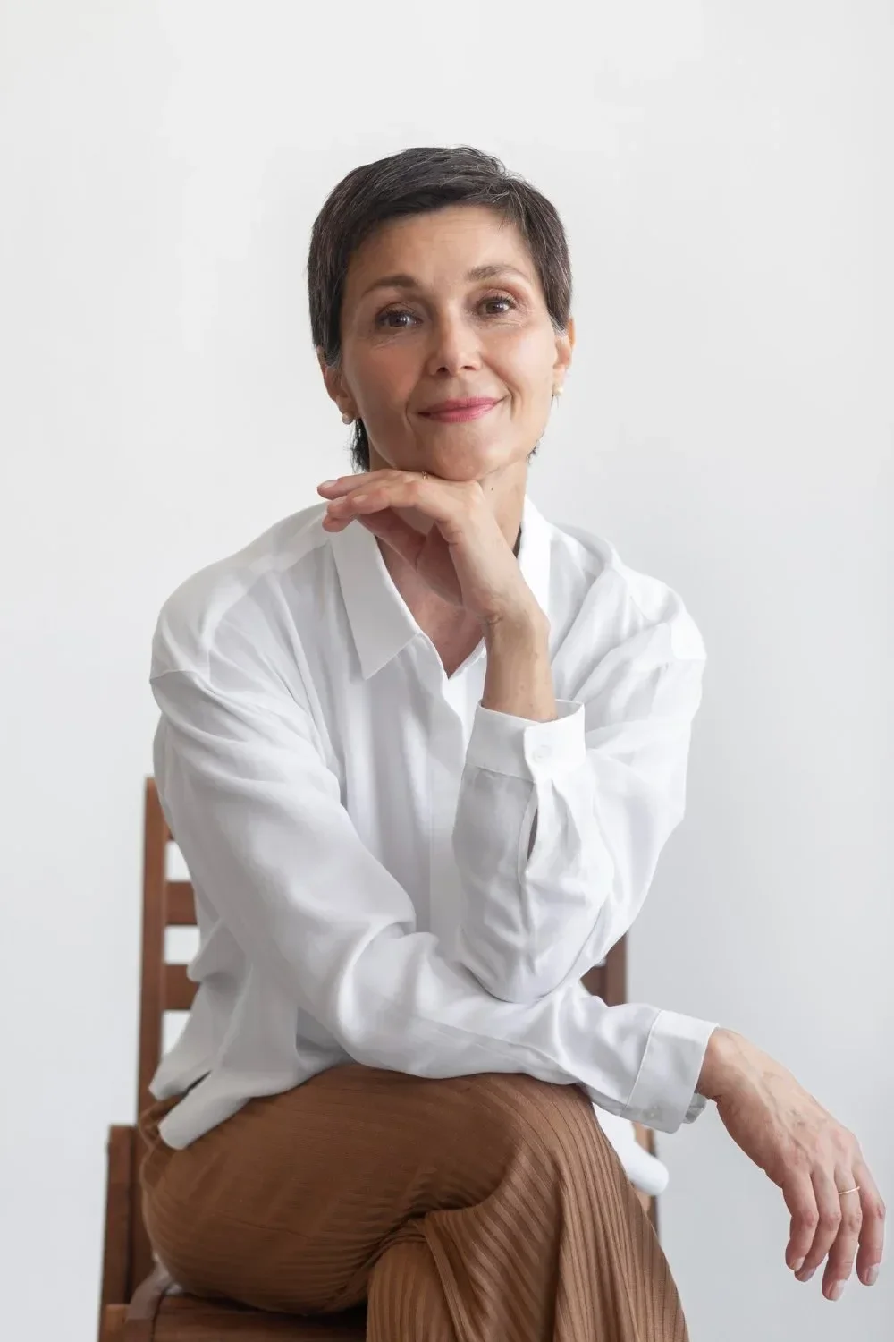 A mature woman with short dark hair wearing a white blouse and brown pants, sitting on a wooden chair with a plain white background, smiling and resting her chin on her hand.