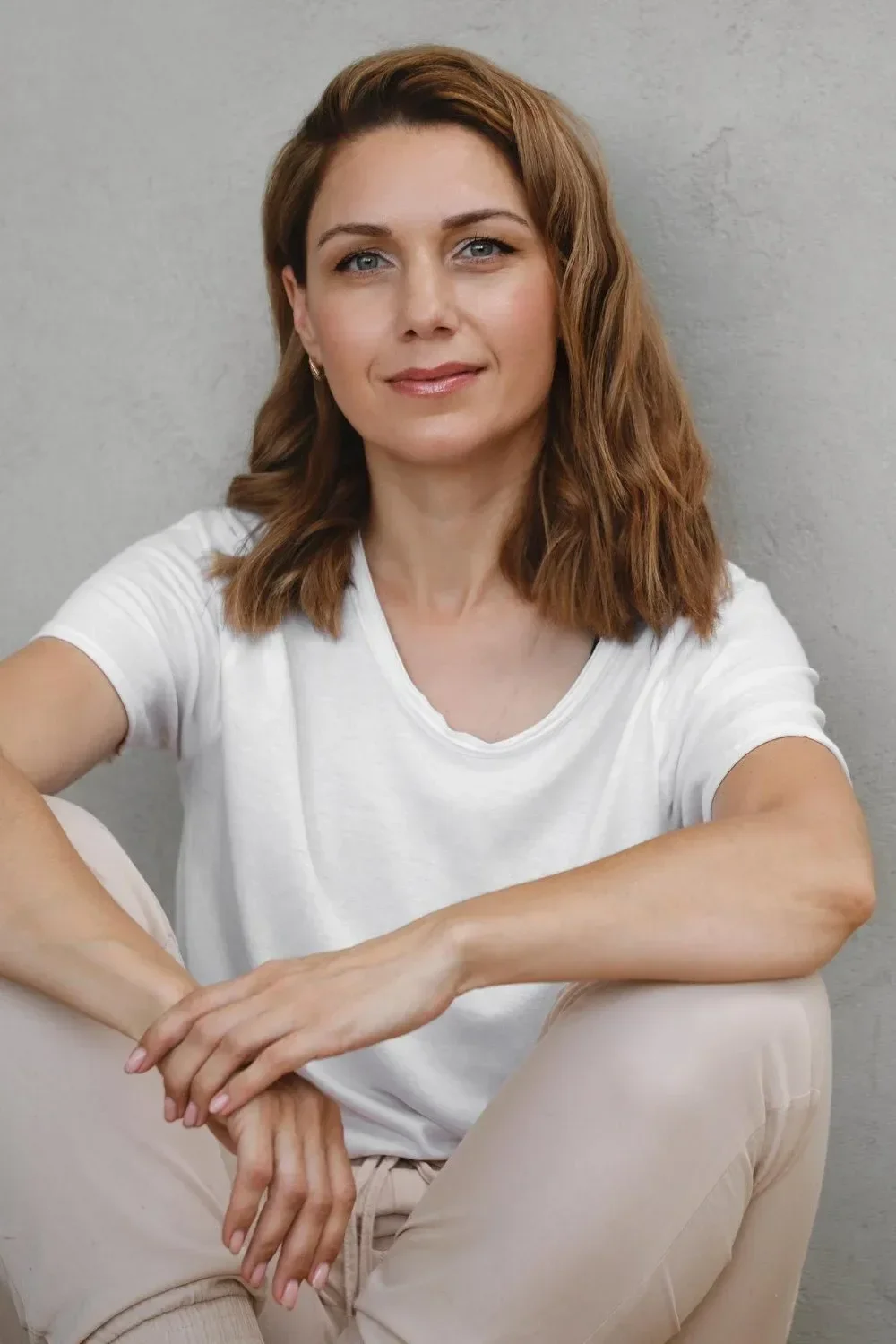 A woman with shoulder-length wavy brown hair and blue eyes, wearing a white t-shirt and beige pants, sitting against a plain gray wall.