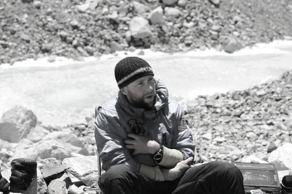 Rob Marshall in outdoor gear sitting on rocks beside a frozen river in a rocky, mountainous area.