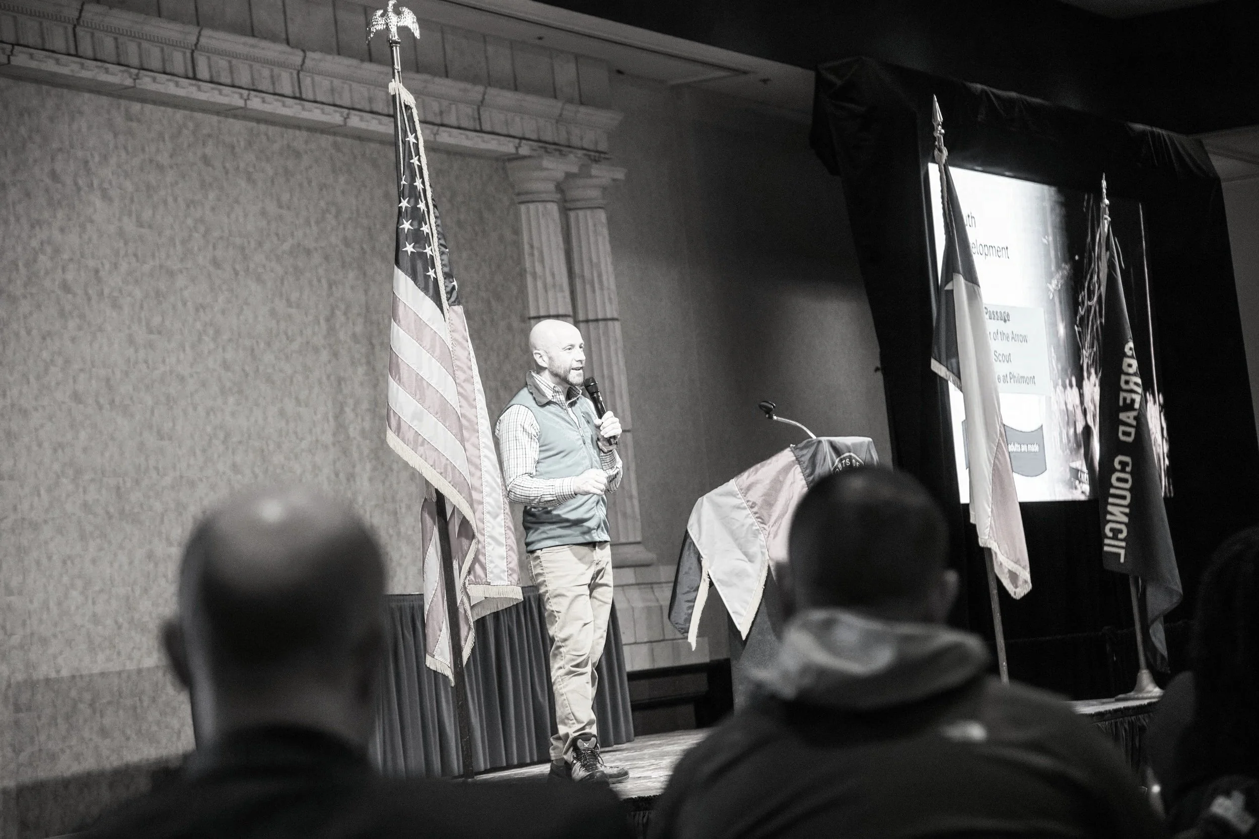 Rob Marshall speaks on stage holding a microphone, giving a presentation in front of an American flag and a screen with a PowerPoint slide, with audience members seated in front of him.