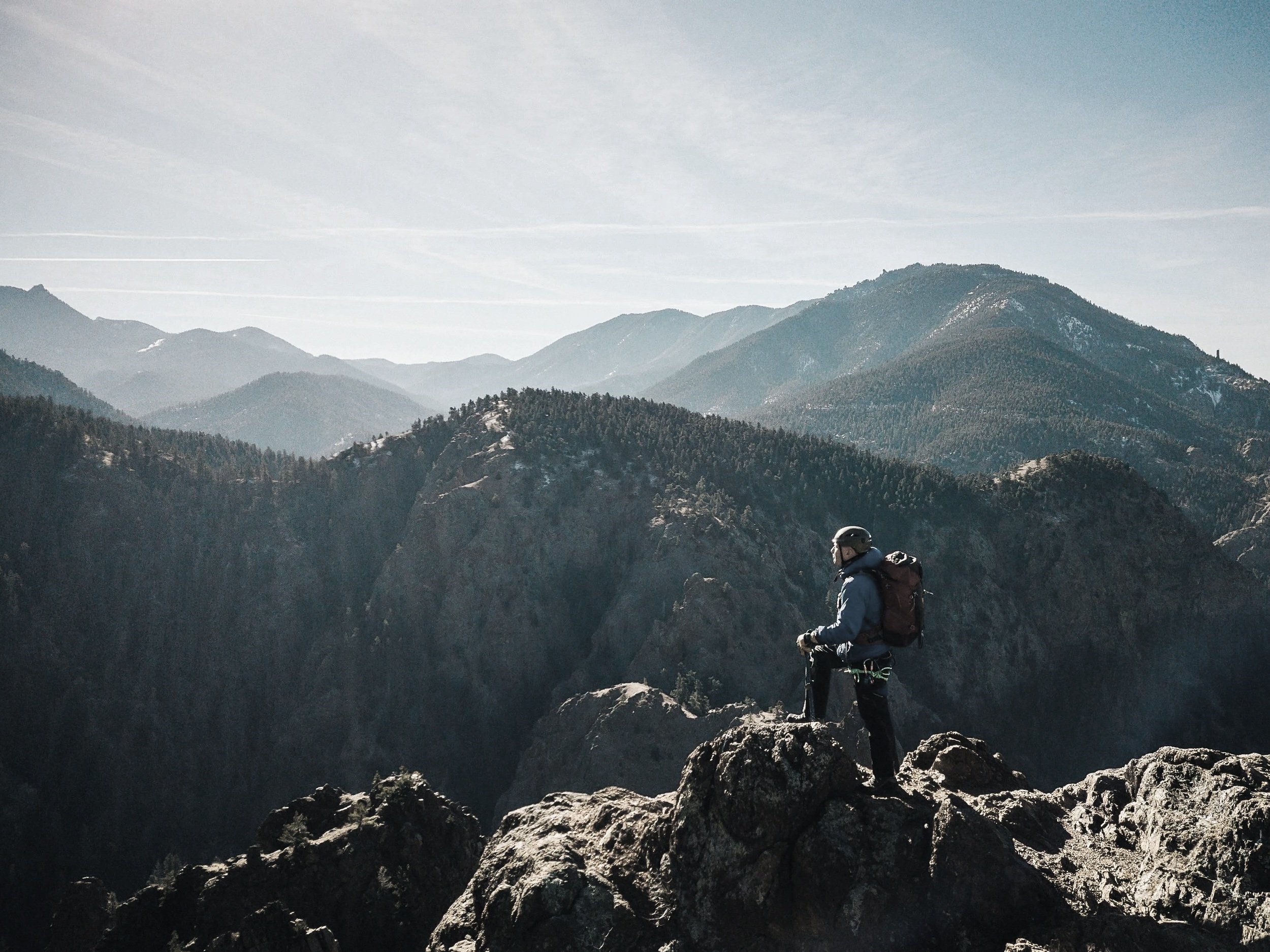 Rob Marshall in outdoor gear, including a helmet and backpack, stands on a large rock overlooking a mountain range with forested hills and a partly cloudy sky.