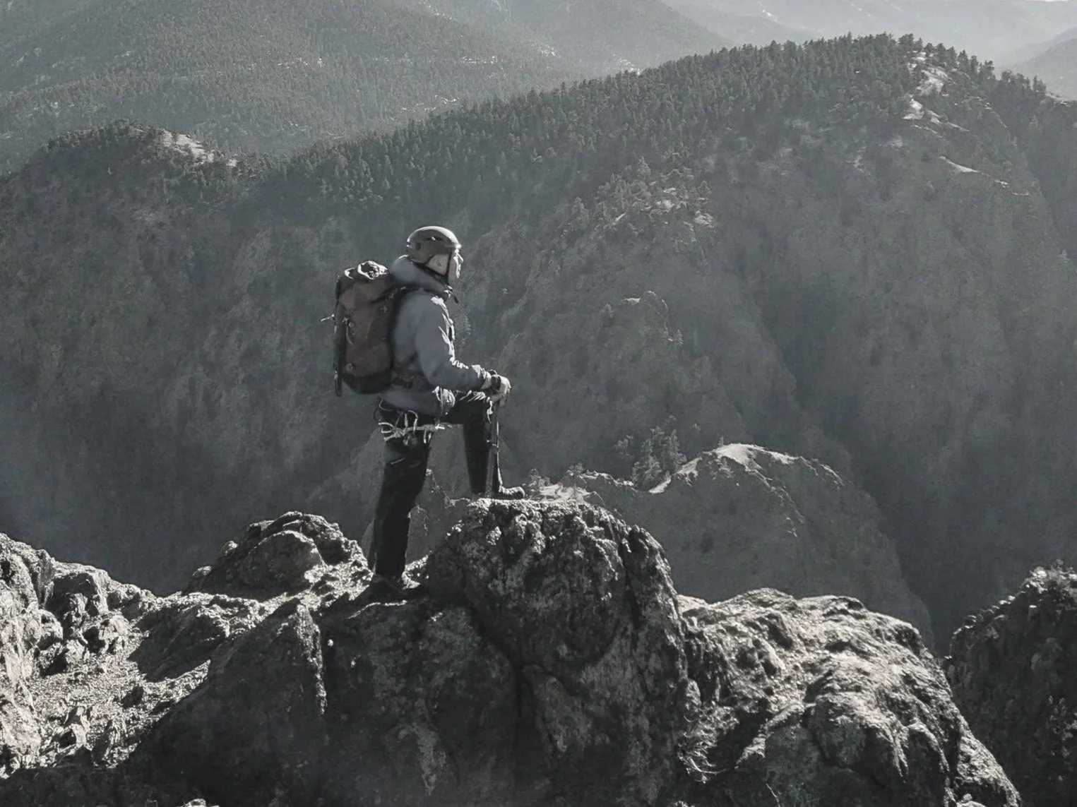 Rob Marshall wearing a helmet and outdoor gear standing on a rocky mountain edge, overlooking a landscape of rugged mountains and forested cliffs in the distance.