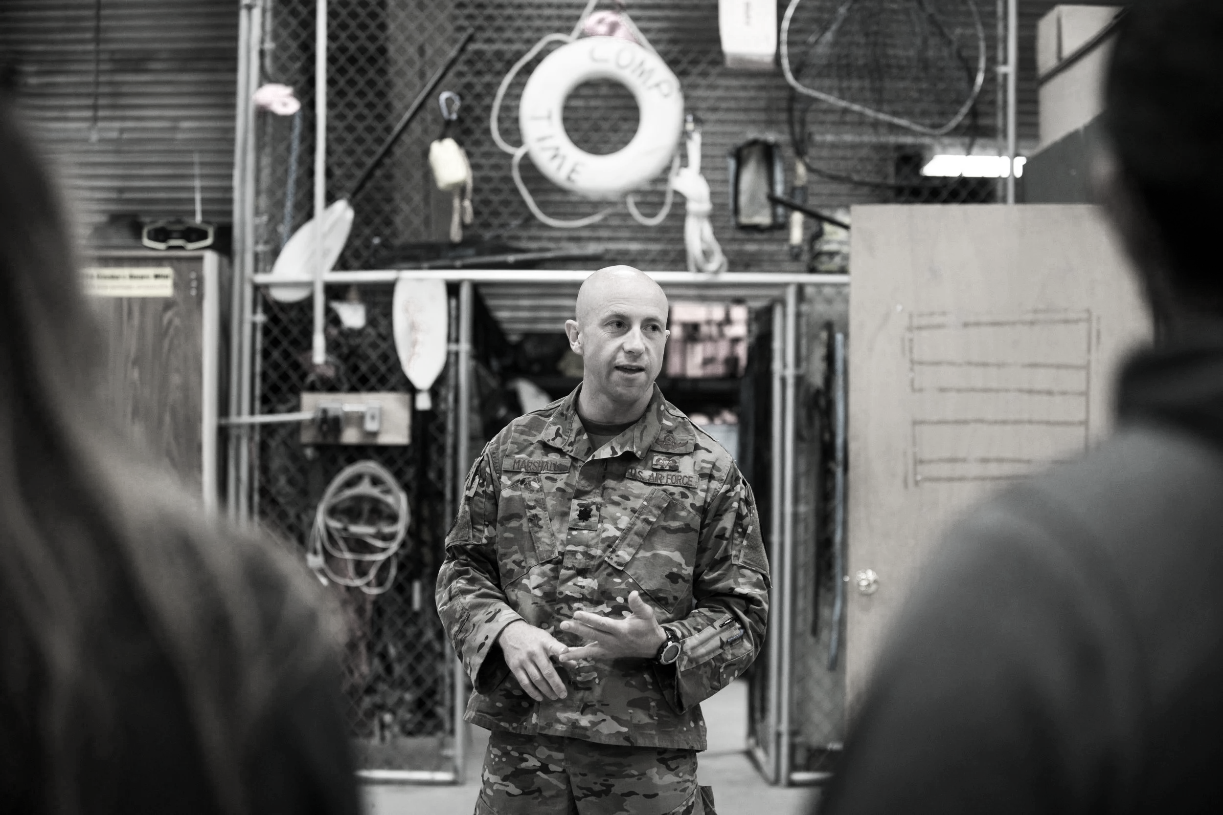 Rob Marshall in military camouflage uniform speaking to a group of people in an industrial or warehouse setting with various equipment and equipment hanging on a wall behind him.