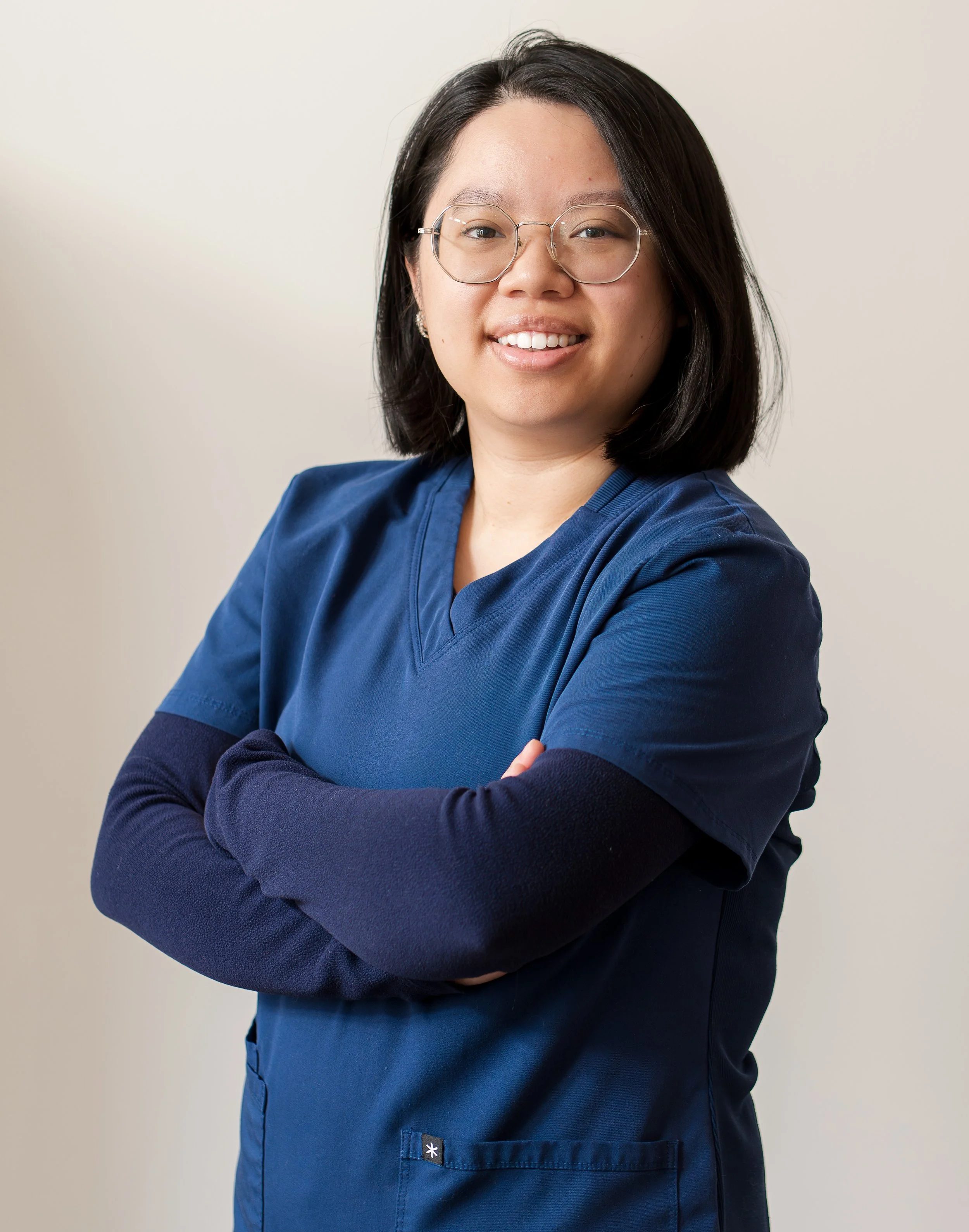 A woman with medium-length black hair, wearing large round glasses, smiling, dressed in blue medical scrubs with her arms crossed, standing against a plain light-colored background.