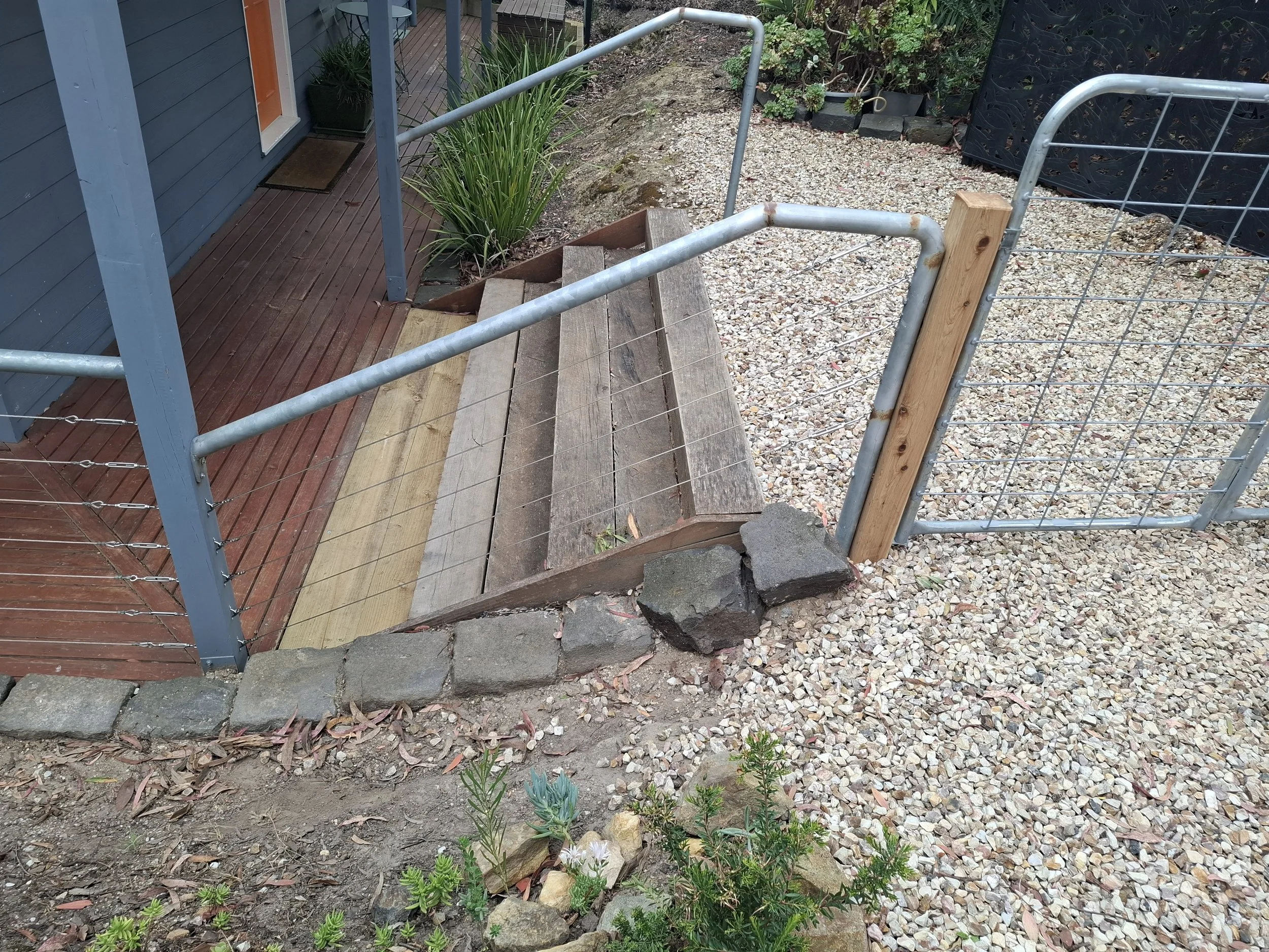 A small outdoor step with a wooden platform, metal railing, and a gravel pathway, next to a blue house with a door, potted plants, and landscaping.