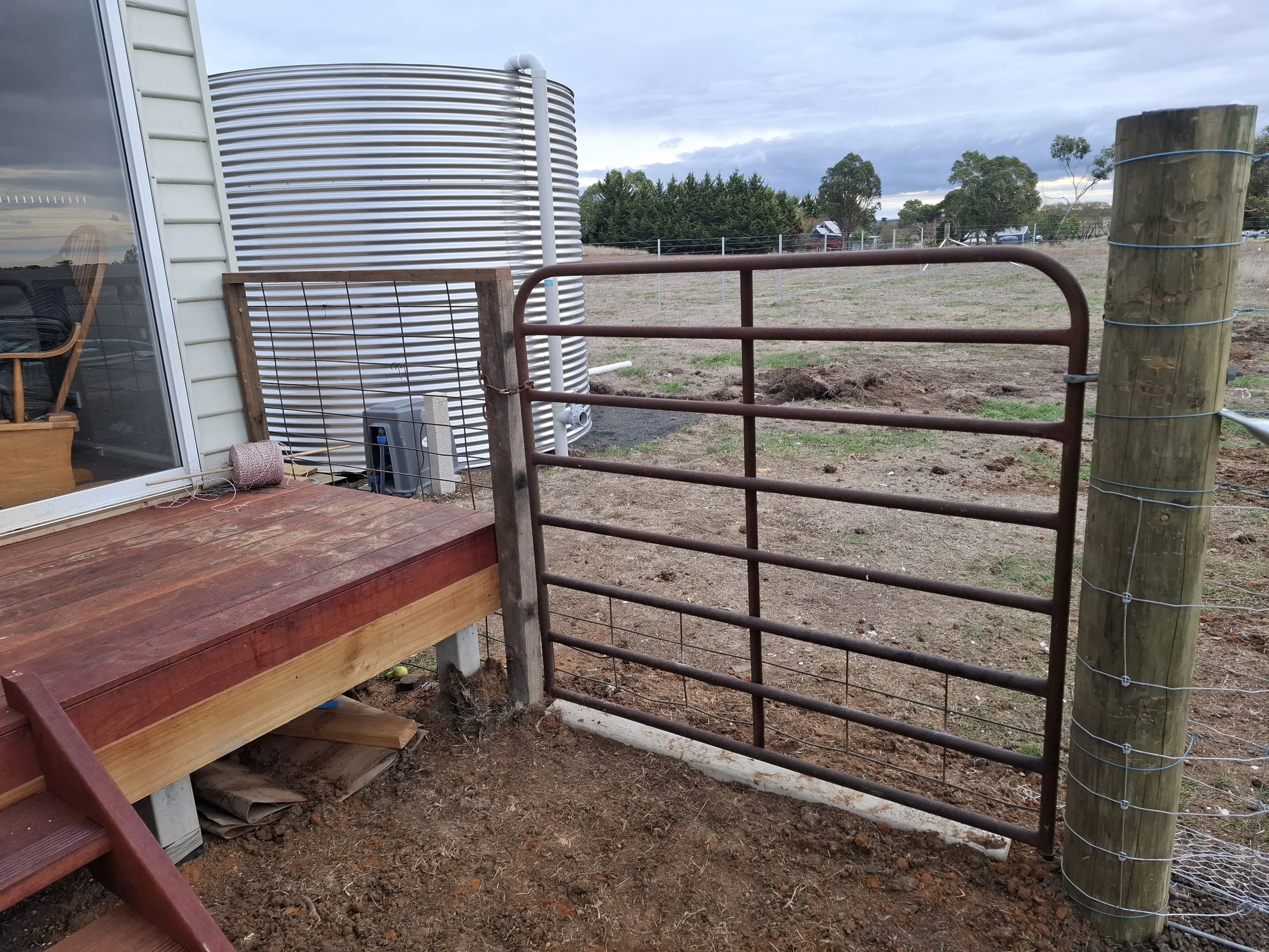 A rural farm scene with a rusty metal gate on a wooden deck, a propane tank, and a large water storage tank in the background, along with open fields and trees under a cloudy sky.