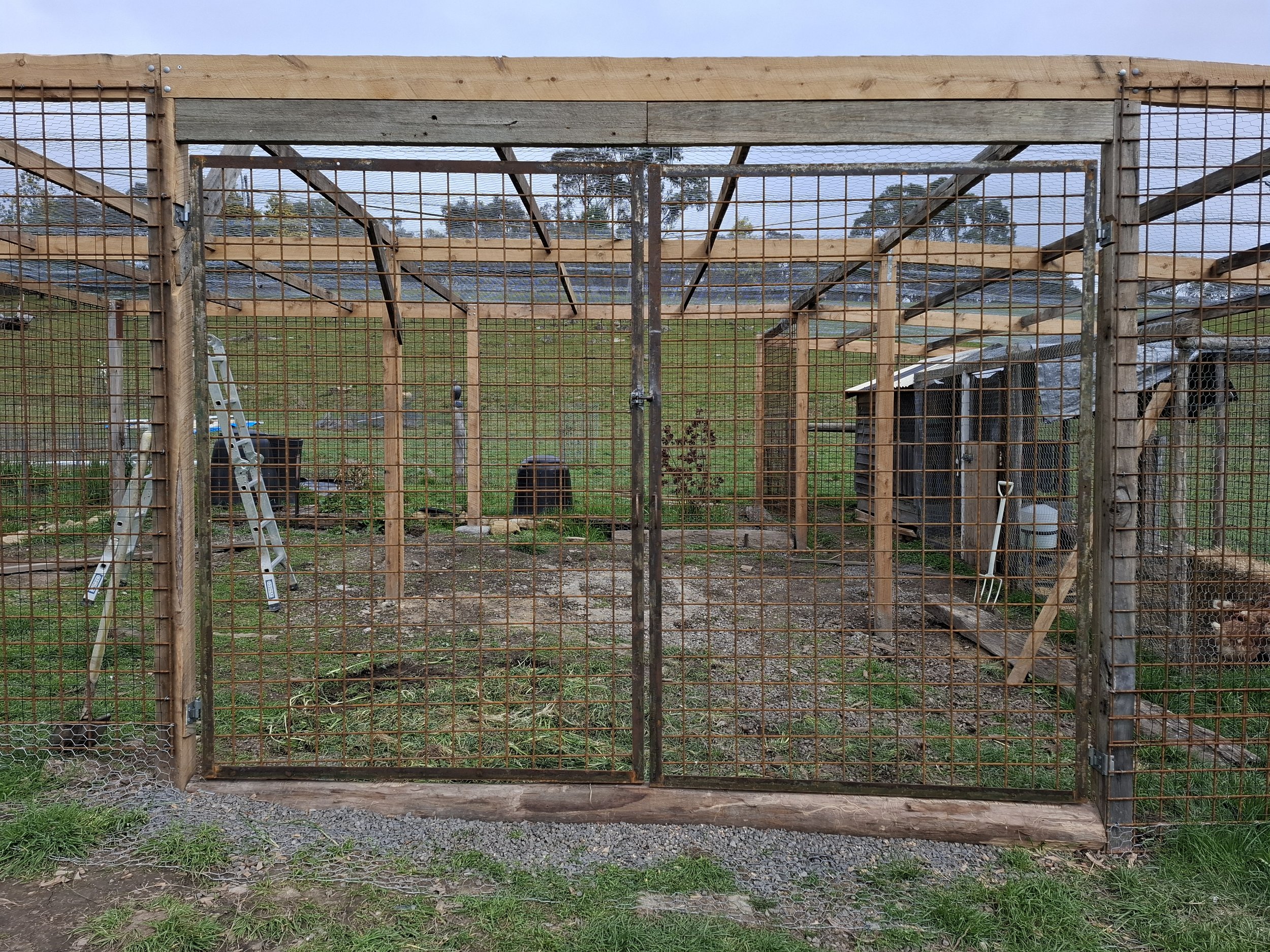A wooden and wire mesh outdoor structure under construction, with a ladder, shovel, and other tools visible, set in a grassy area with hills and trees in the background.