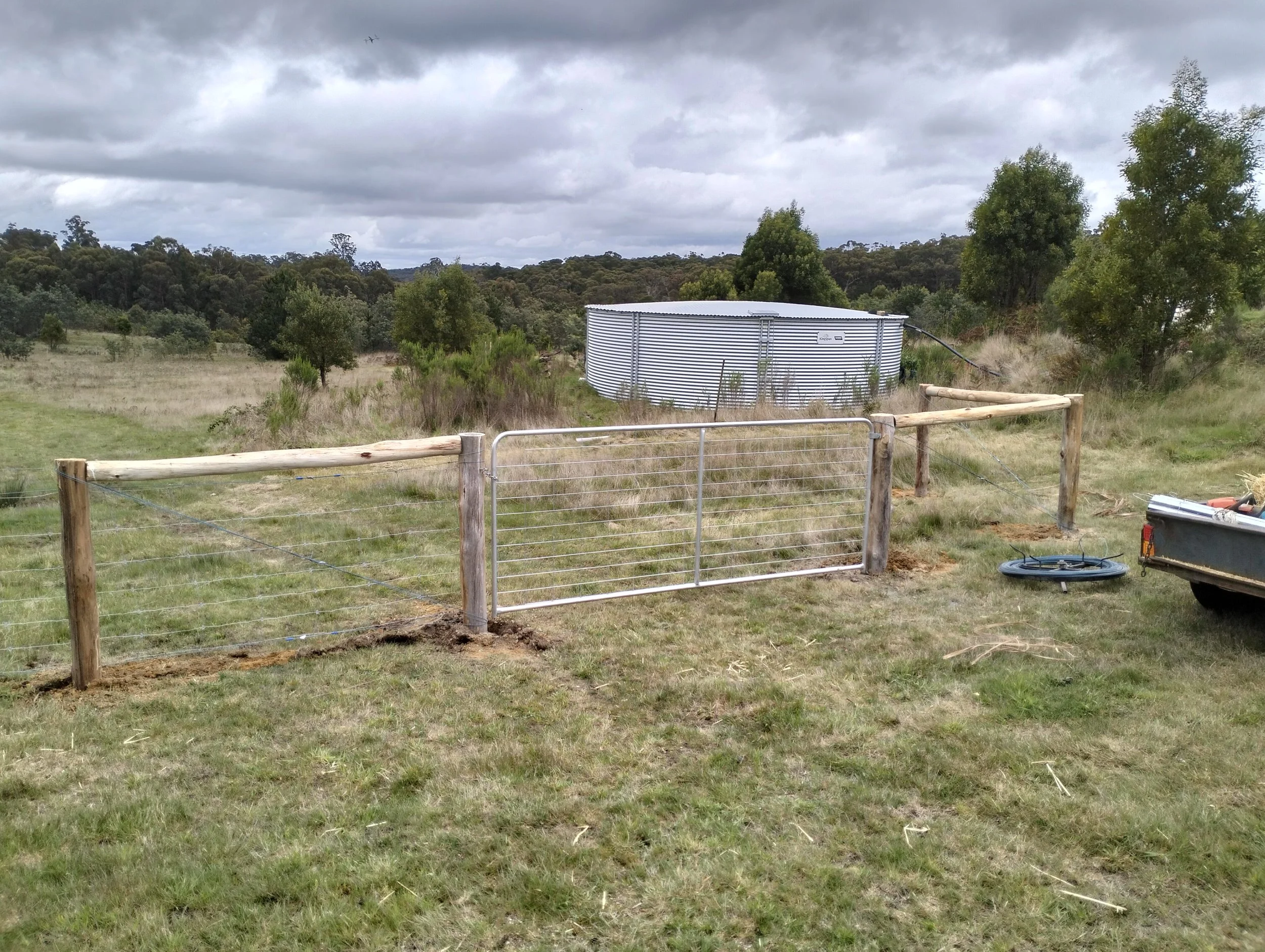 A fenced field with a metal gate, a corrugated metal water tank, trees, and cloudy sky.