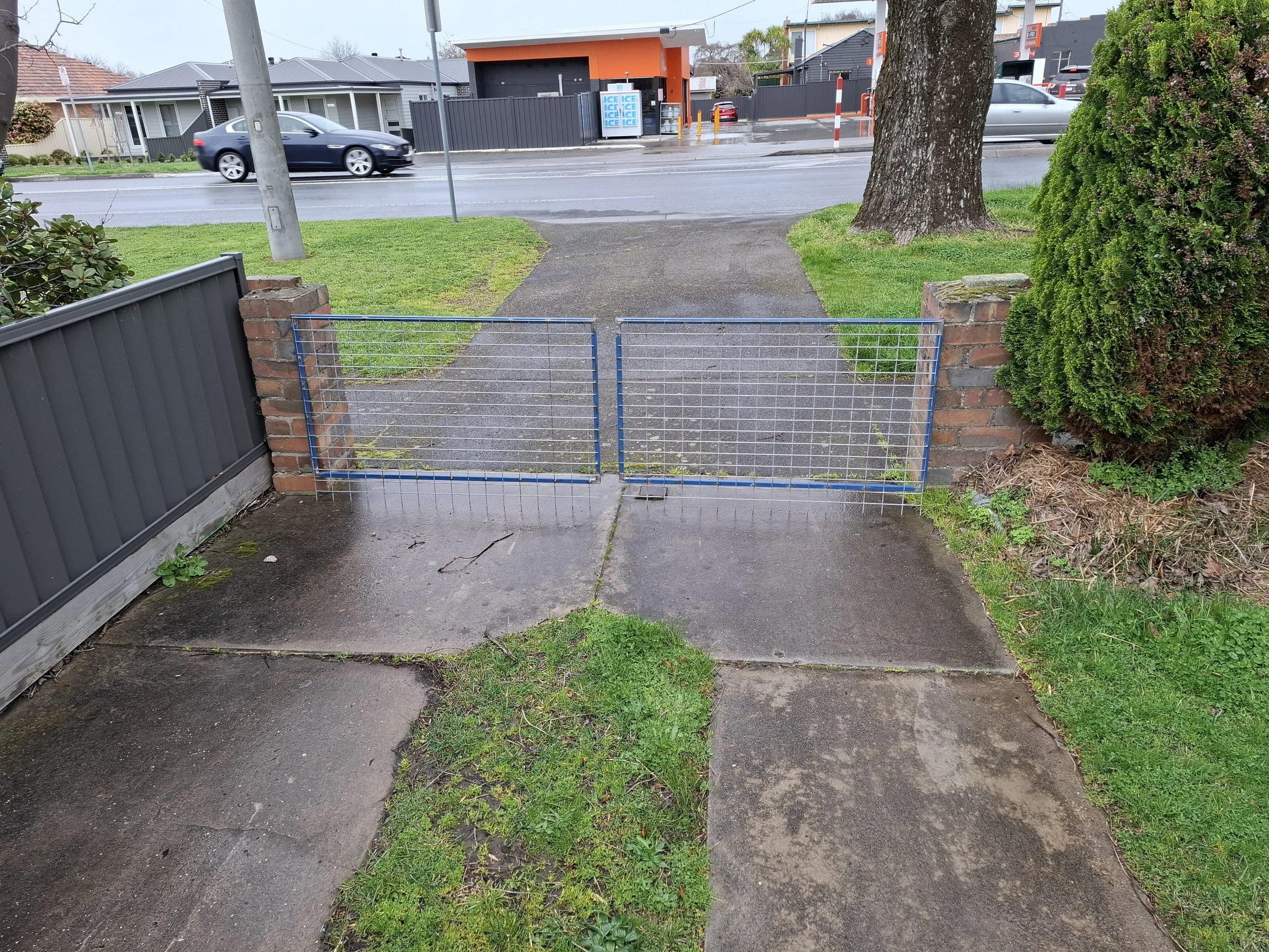 A small gate blocking the sidewalk leading to the street, with a brick wall and a fence on the left, and a brick pillar and bush on the right. A tree is further back on the right, and the street outside has cars passing. In the background, there is a