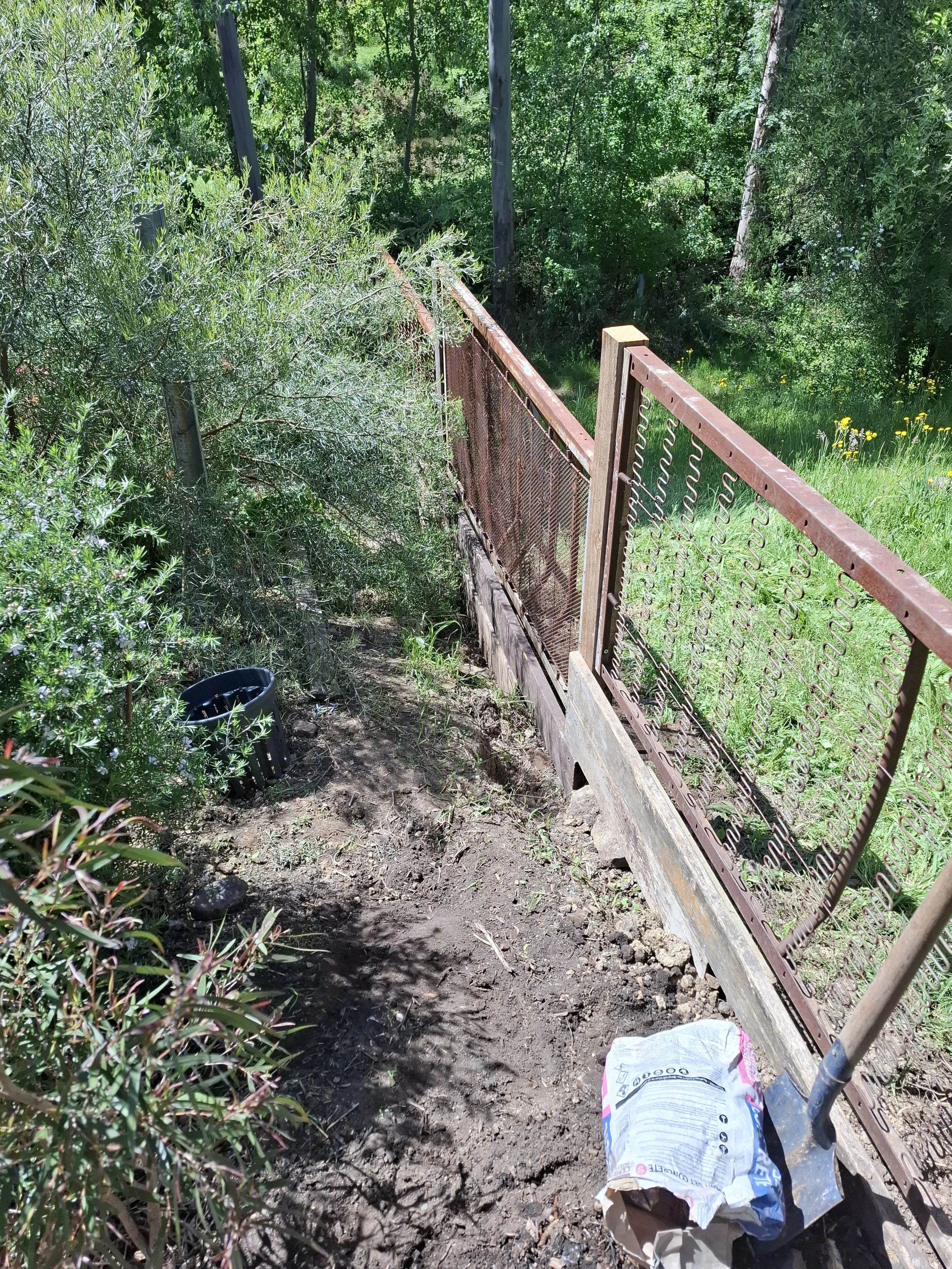 A narrow outdoor pathway with a dirt surface, bordered by green bushes and trees, with a rusted iron fence on the right side and gardening tools, including a shovel and a bag of soil or mulch, placed on the ground.