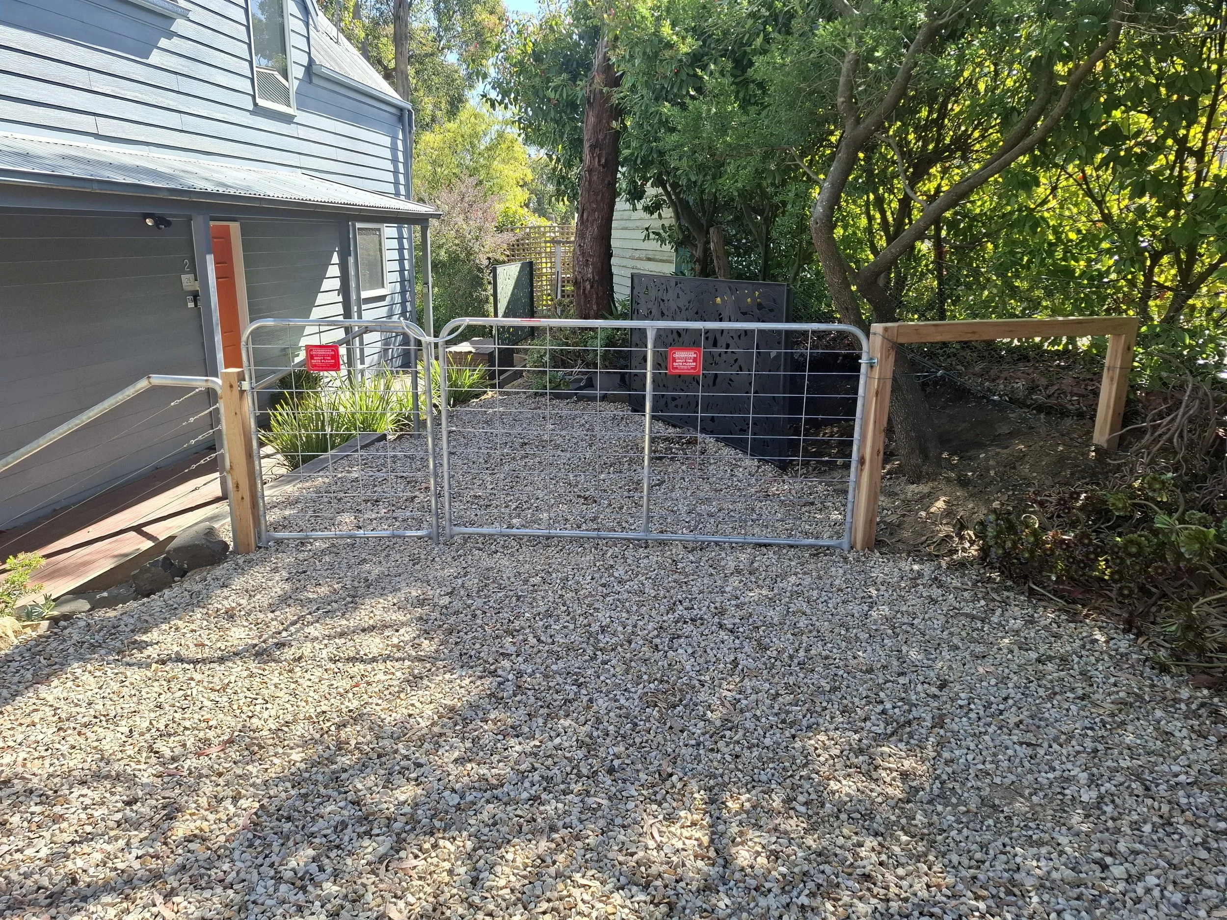 A small fenced area with gravel ground, metal gate, wooden posts, and surrounding greenery including trees and plants in a backyard. There is a gray house on the left with a door and window, and a black decorative panel in the background.