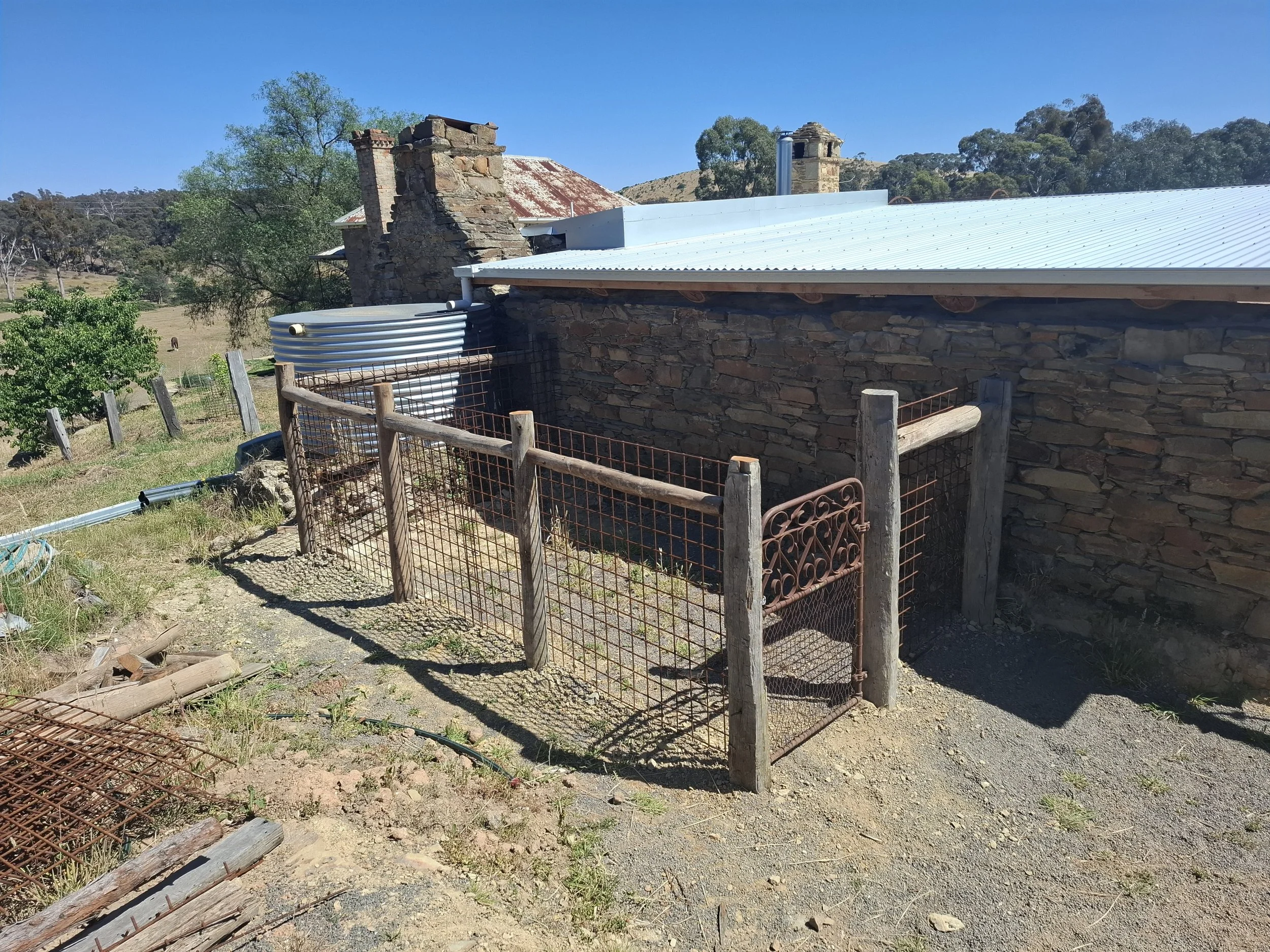 A rustic outdoor scene featuring a stone building with a metal roof, surrounded by a simple wood and metal fence, in a rural landscape with trees and distant hills under a clear blue sky.