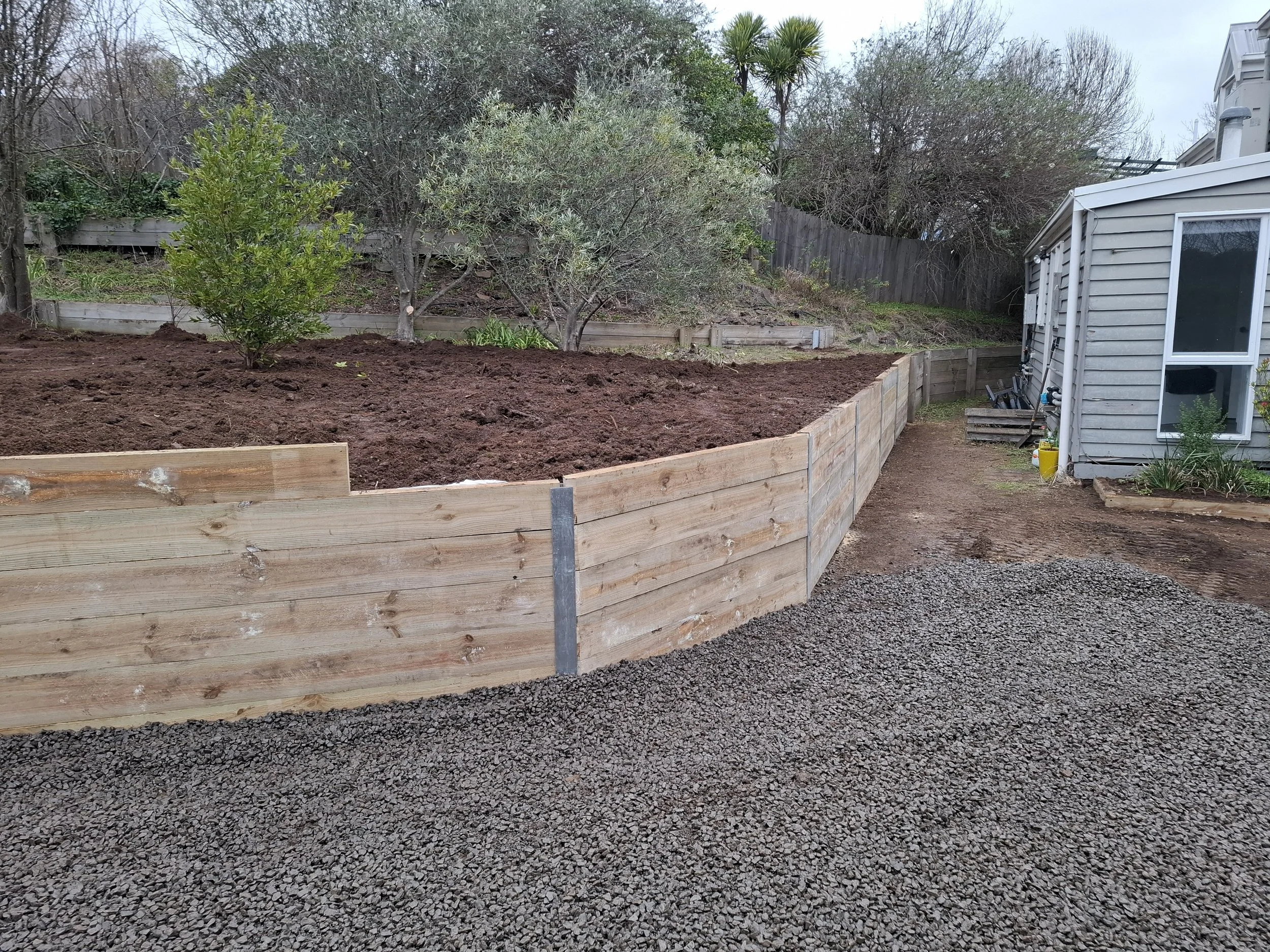 A backyard with a newly built wooden retaining wall, soil, and a small garden area with trees and plants near a house.