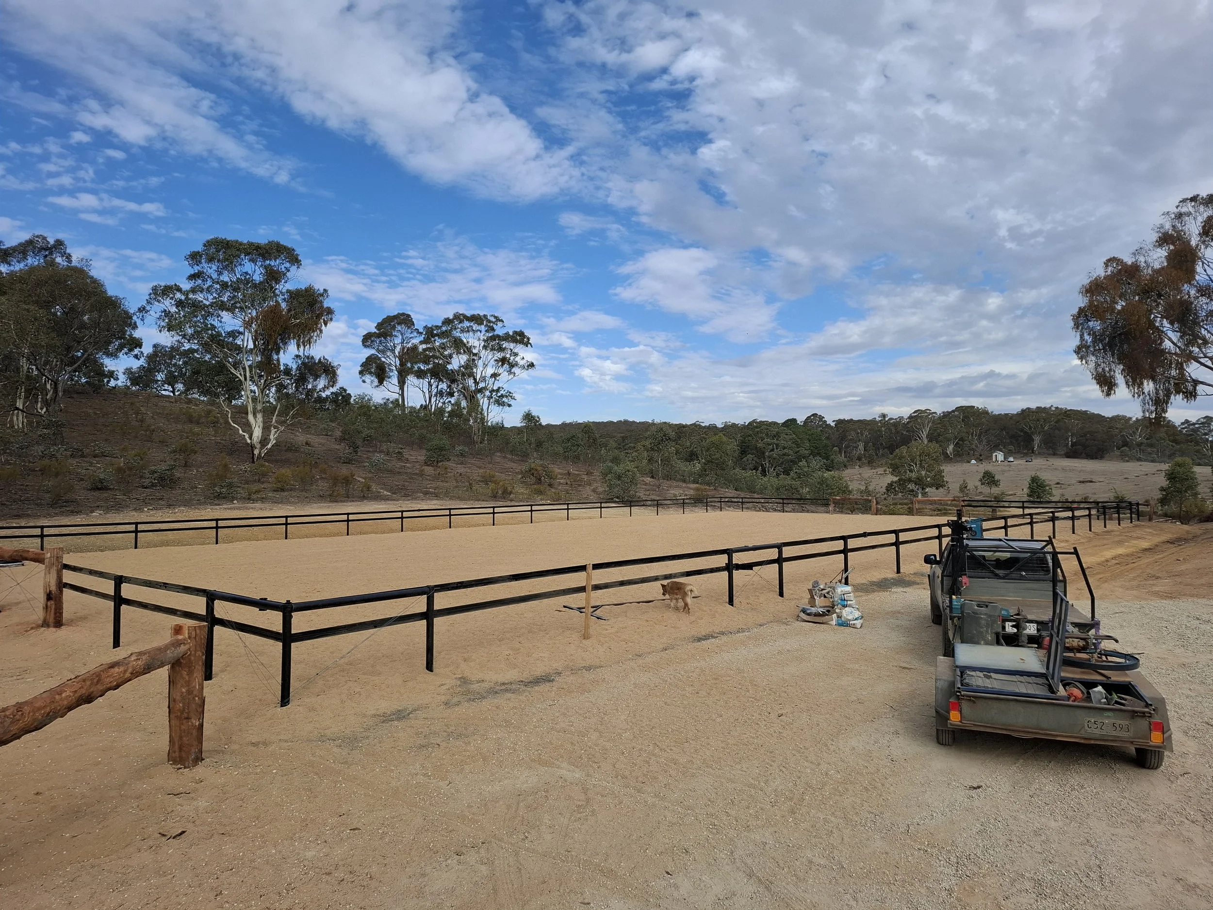 An outdoor riding arena with sandy footing, surrounded by a black wooden fence. In the background, there are trees and a blue sky with scattered clouds. Near the fence, there is a small utility vehicle and some supplies on the ground.
