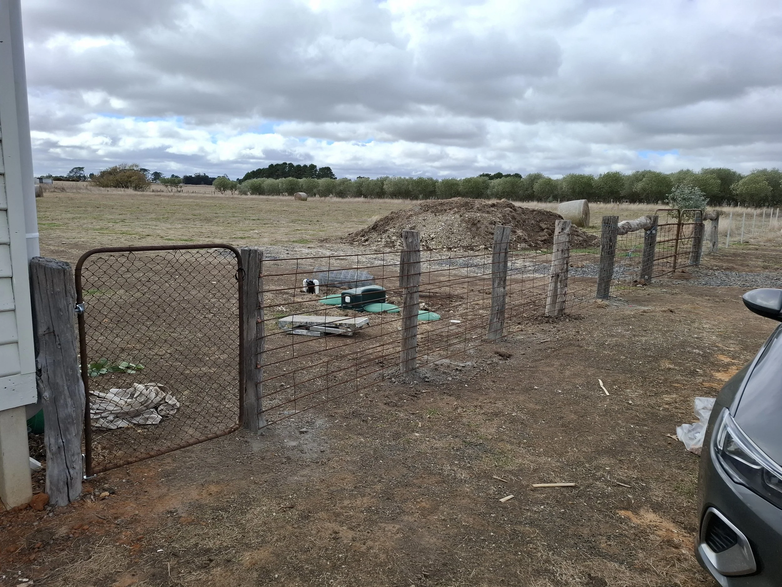 A rural farmyard with a wire gate, a dirt pathway, and a fenced field with hay bales and trees in the background. Overcast sky.