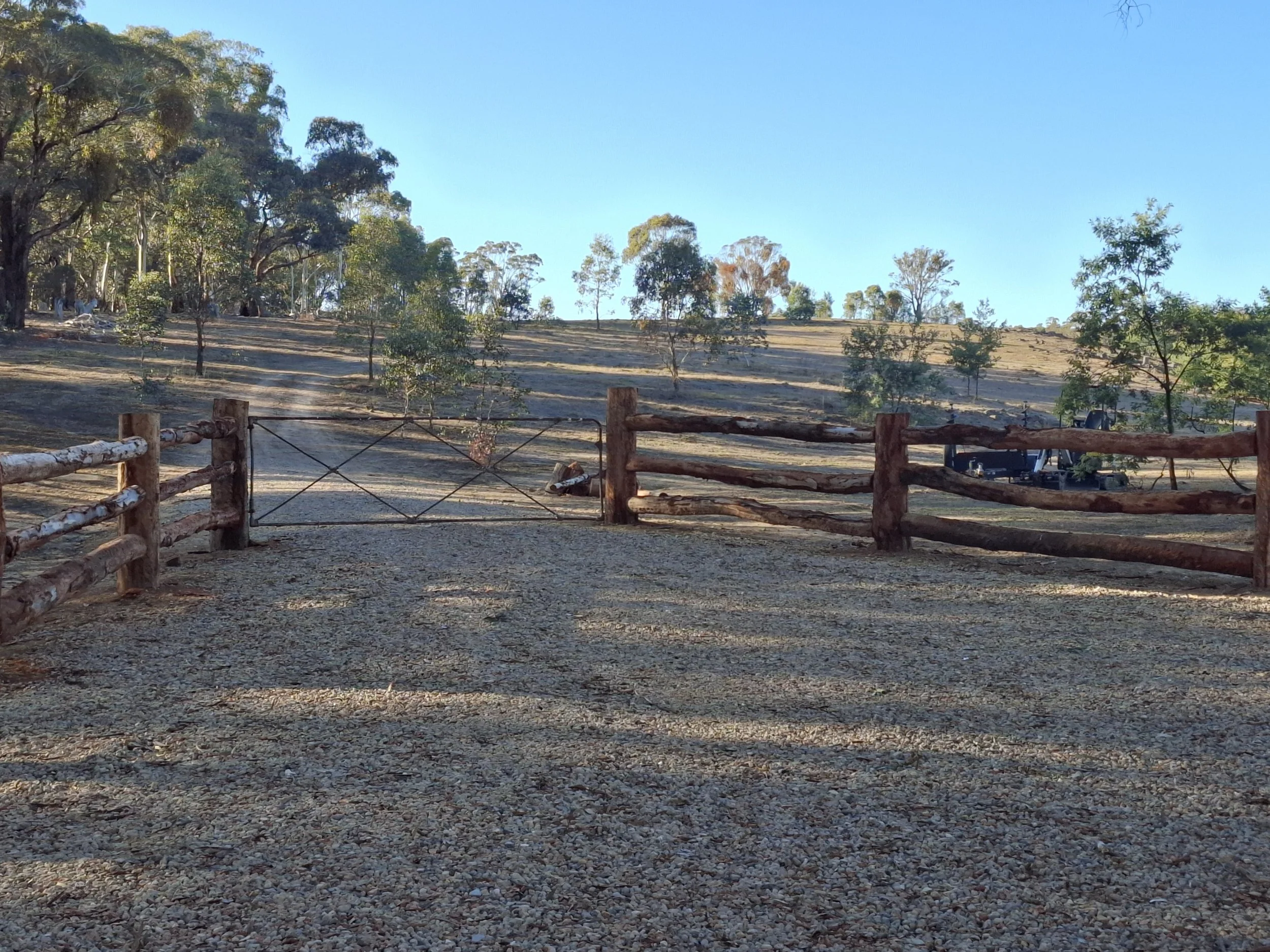 A gravel pathway with wooden fences on either side, leading to a hill with sparse trees and a clear blue sky in the background.