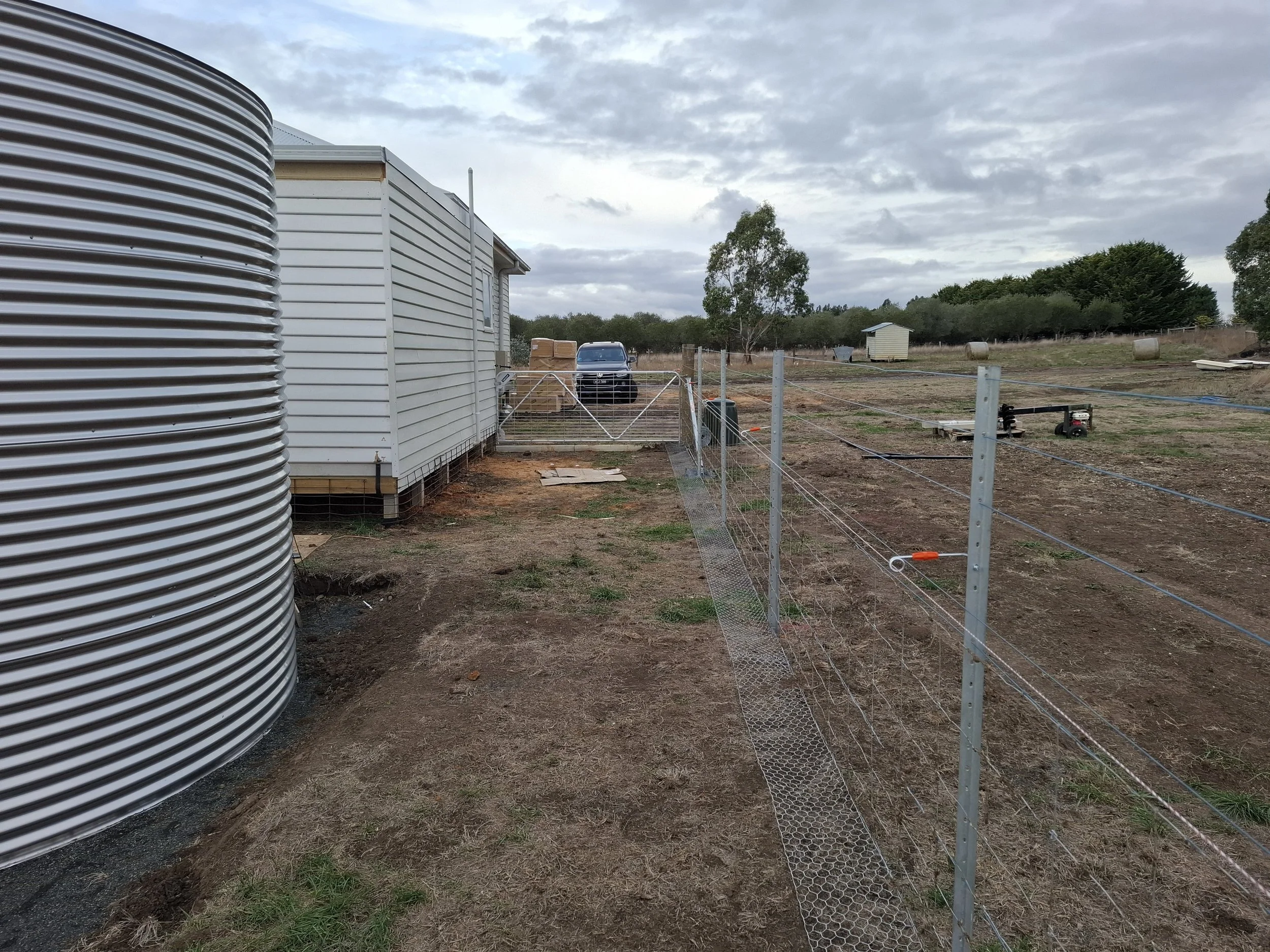 A rural farm scene with a white mobile home, a large metal water tank on the left, a wire fence, and a black truck parked in the background among trees and open land.