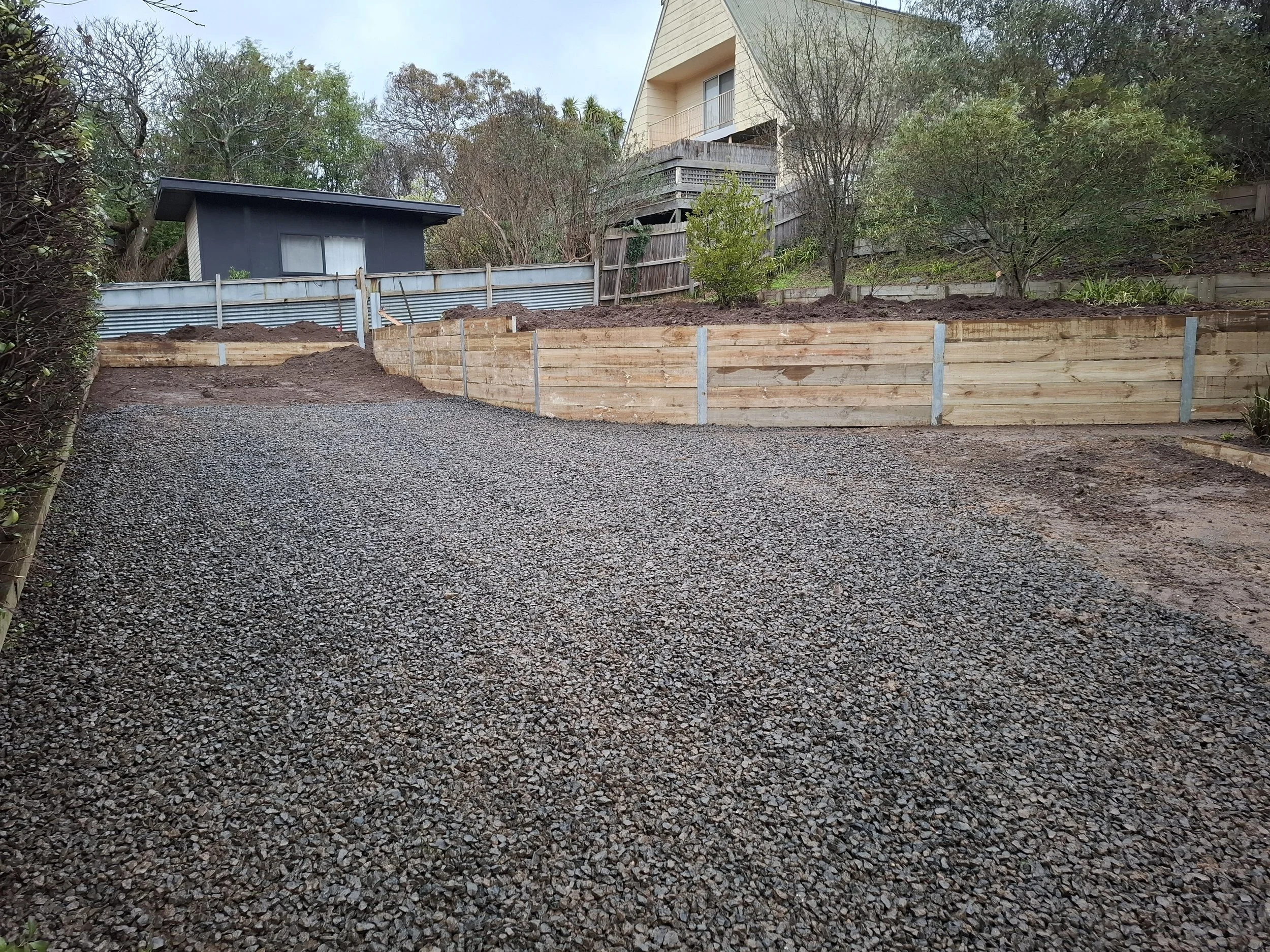 Newly graded driveway with gravel, wooden retaining wall, and some trees and houses in the background.