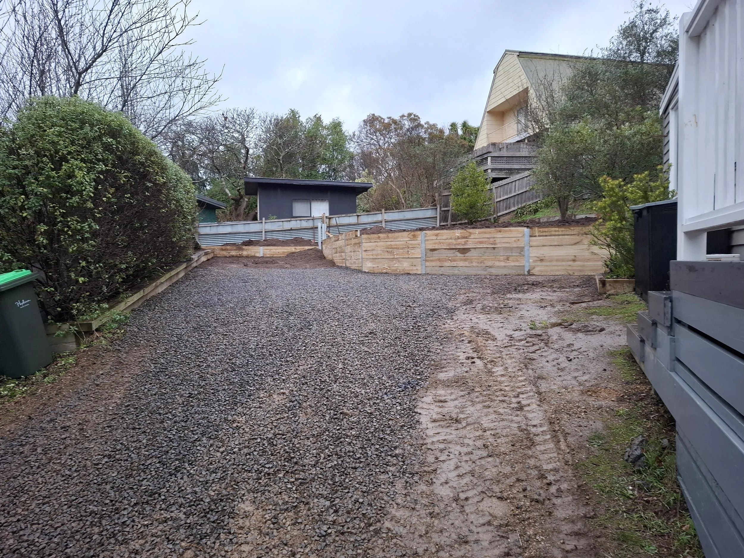 A gravel driveway with a wooden retaining wall on the right side, leading up to a small black shed at the top, with trees and a house in the background.