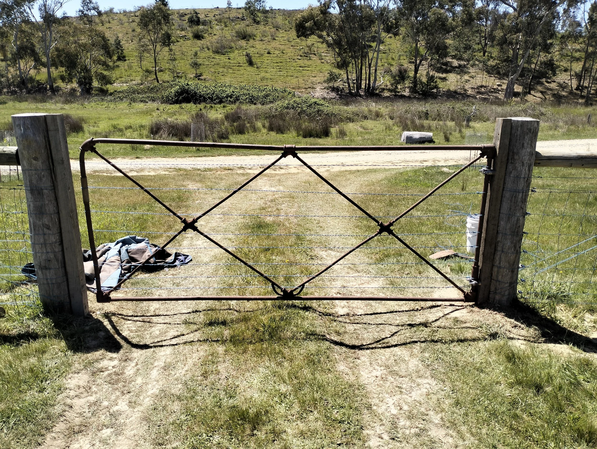 A rusty metal gate in front of a grassy field with a dirt road, trees, and hills in the background.