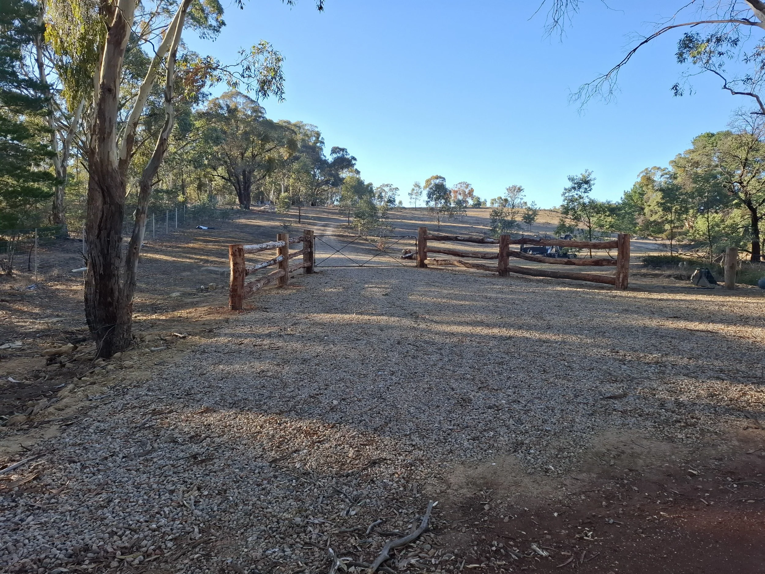 A gravel driveway leading to a wooden gate in a rural area. Trees and a clear blue sky are visible in the background.
