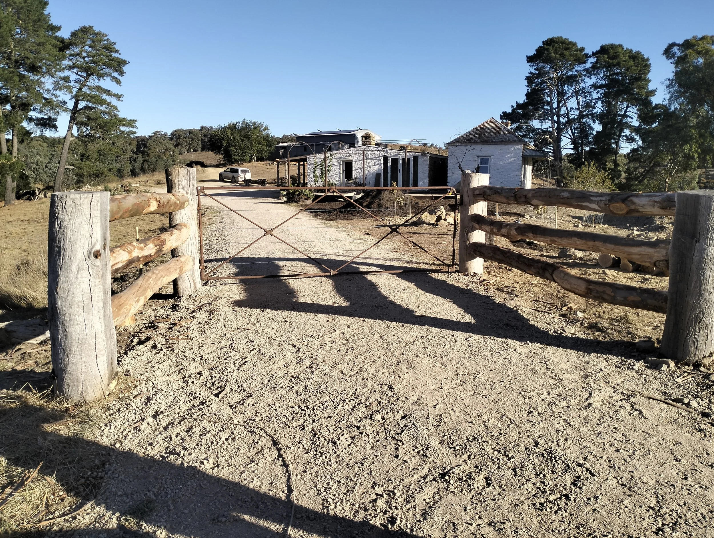 A gravel driveway with a rusty metal gate, flanked by wooden posts, leading to a white house with a porch. A car is parked near the house, surrounded by trees and open land under a clear blue sky.
