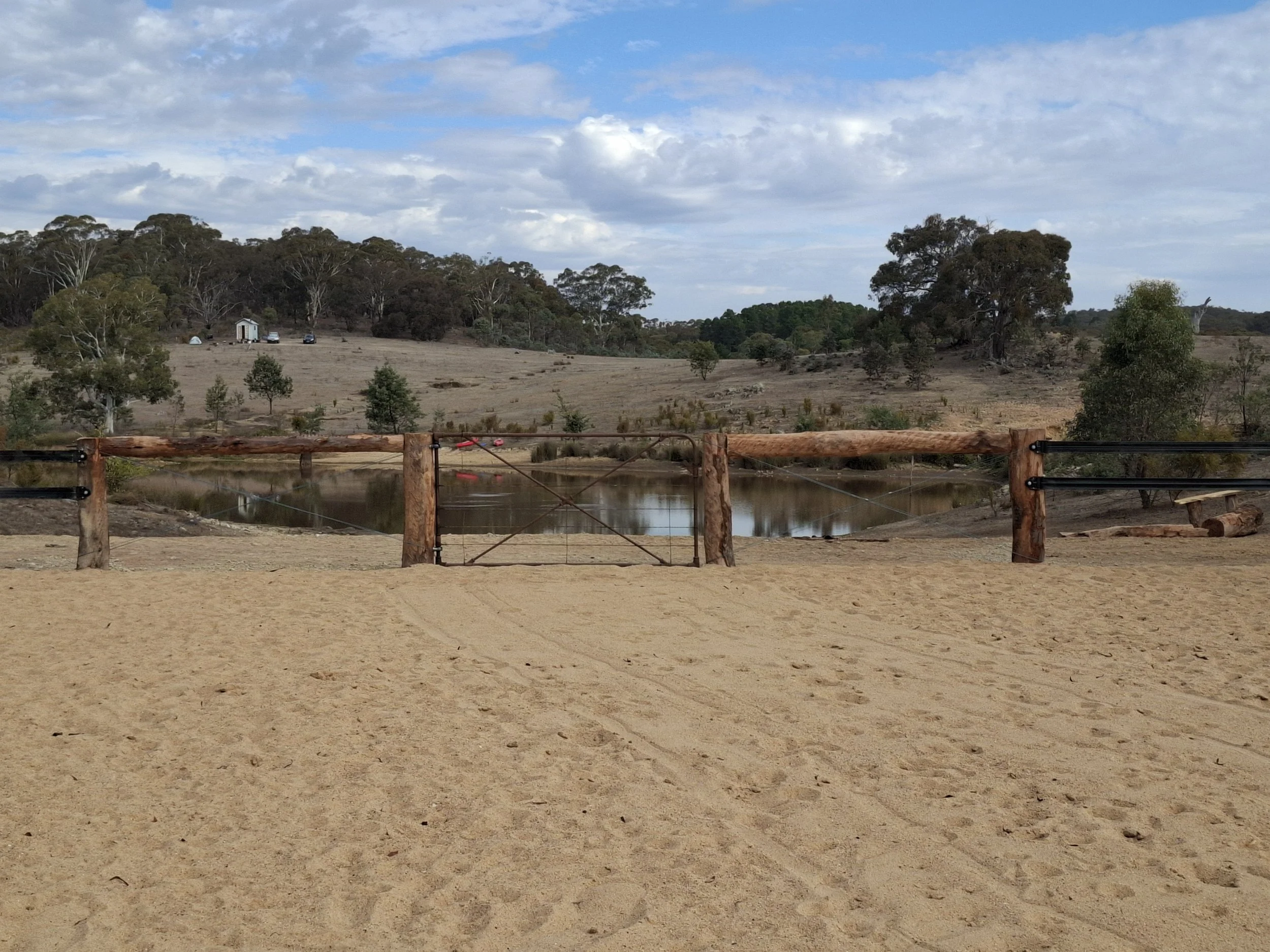 A rustic wooden fence with a metal gate separates a sandy area from a small lake or pond. In the background, there are rolling hills with scattered trees and a few small buildings or sheds. The sky is partly cloudy.