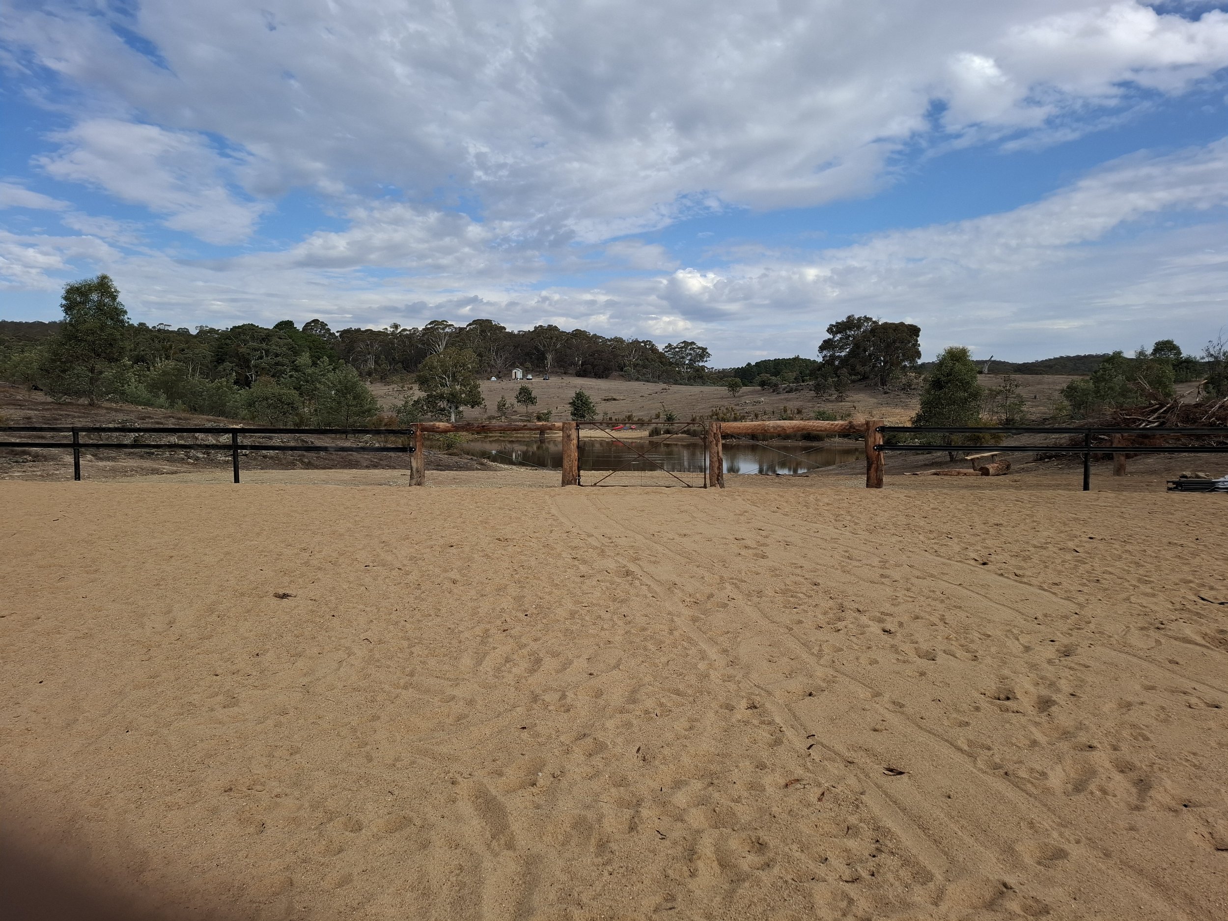 View of a sandy area with a wooden and metal fence, a water body, trees, and rolling hills under a partly cloudy sky.