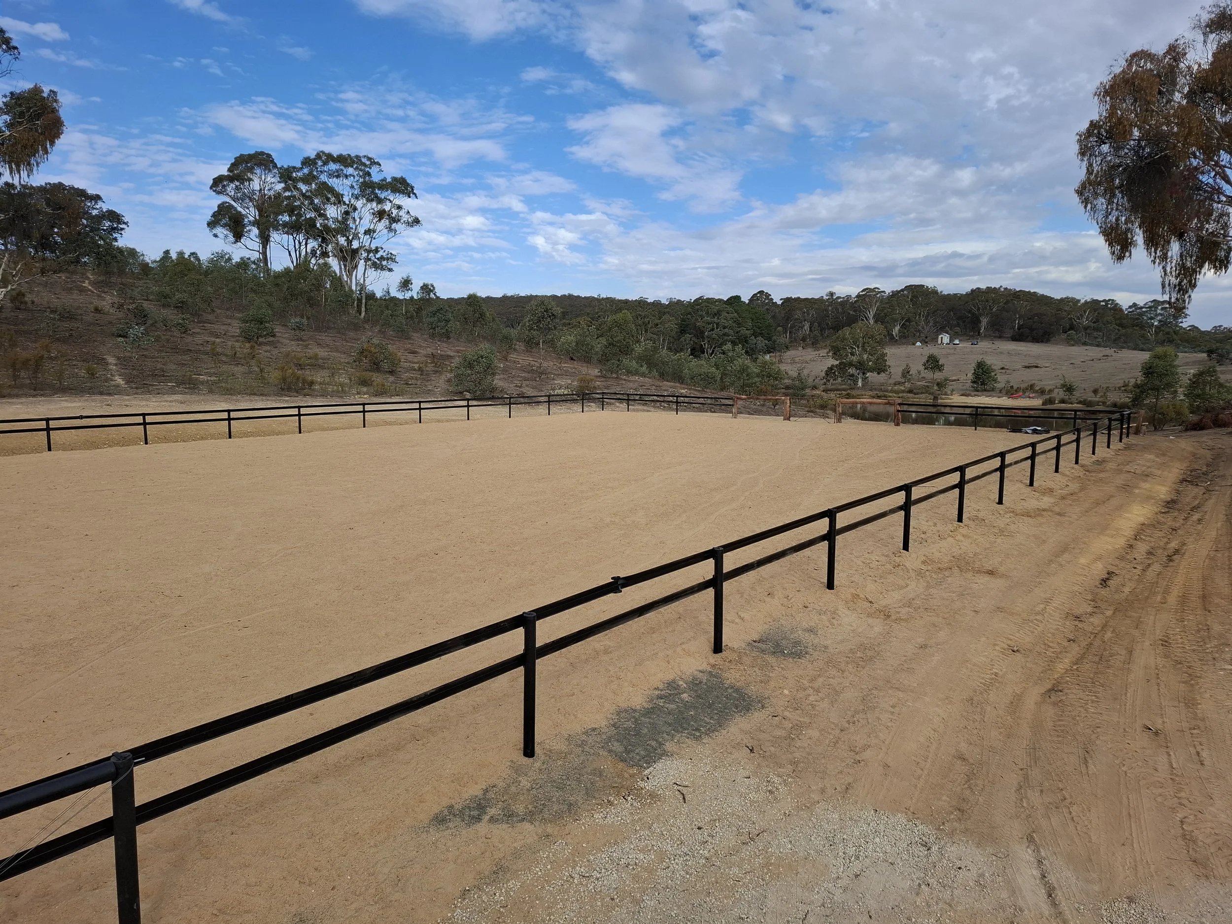 A fenced outdoor riding arena with a dirt and sand surface, surrounded by trees and rolling hills under a partly cloudy sky.