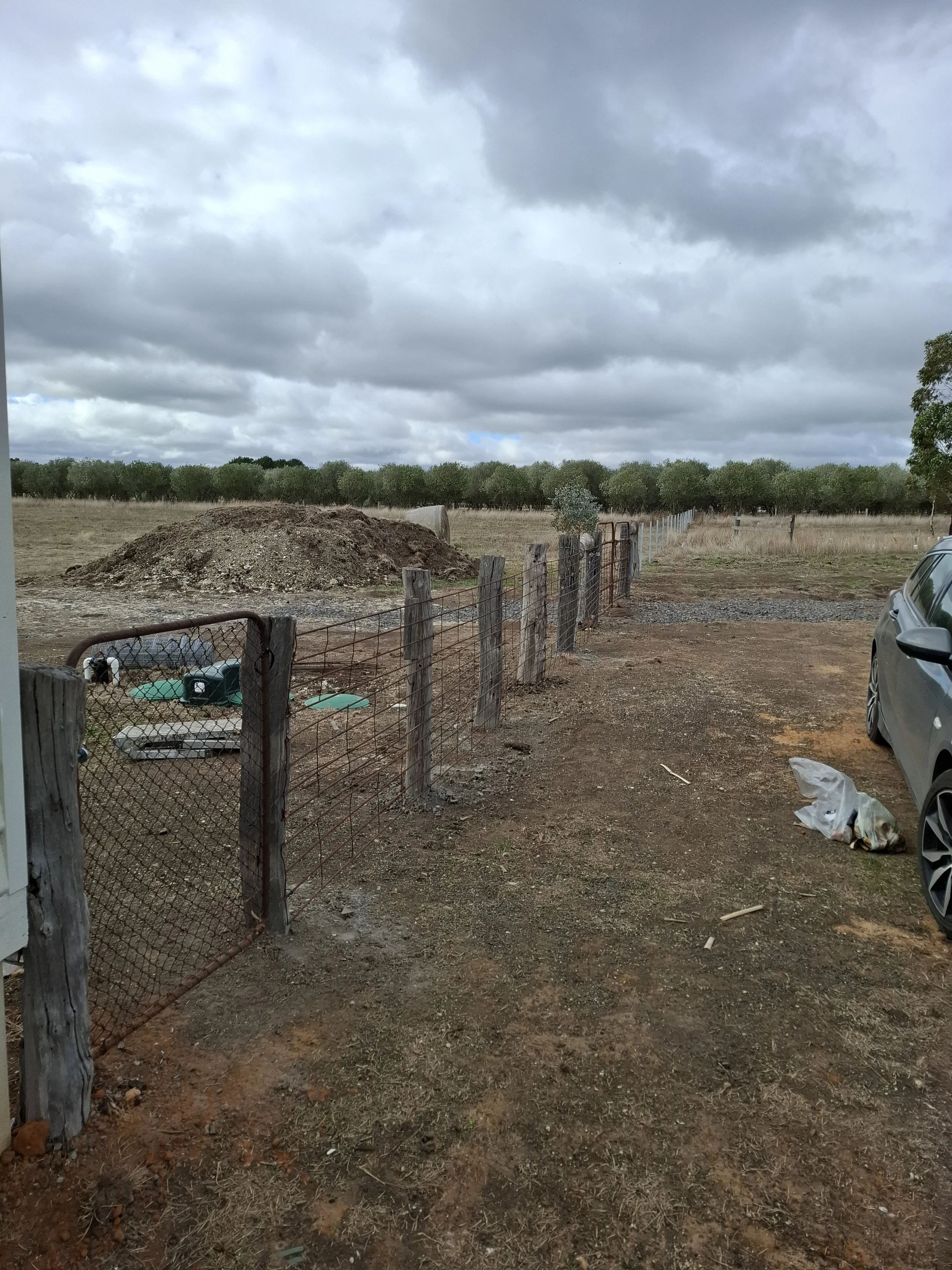 A dirt driveway next to a metal and wooden fence leading to a field with trees in the background. Overcast sky with gray clouds.