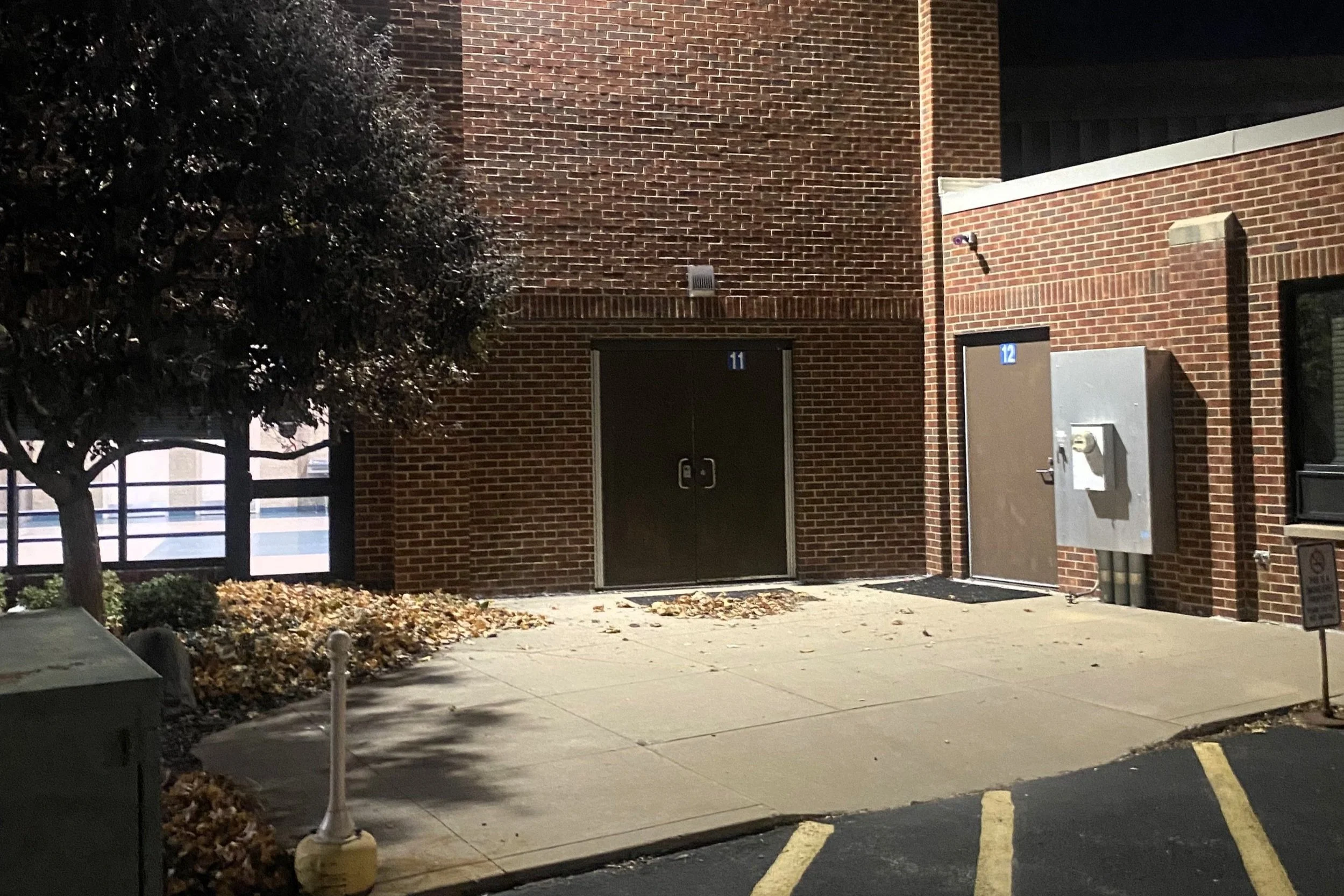 Night view of a brick building with two doors numbered 11 and 12, a small window, a tree, and parking spaces with yellow lines.