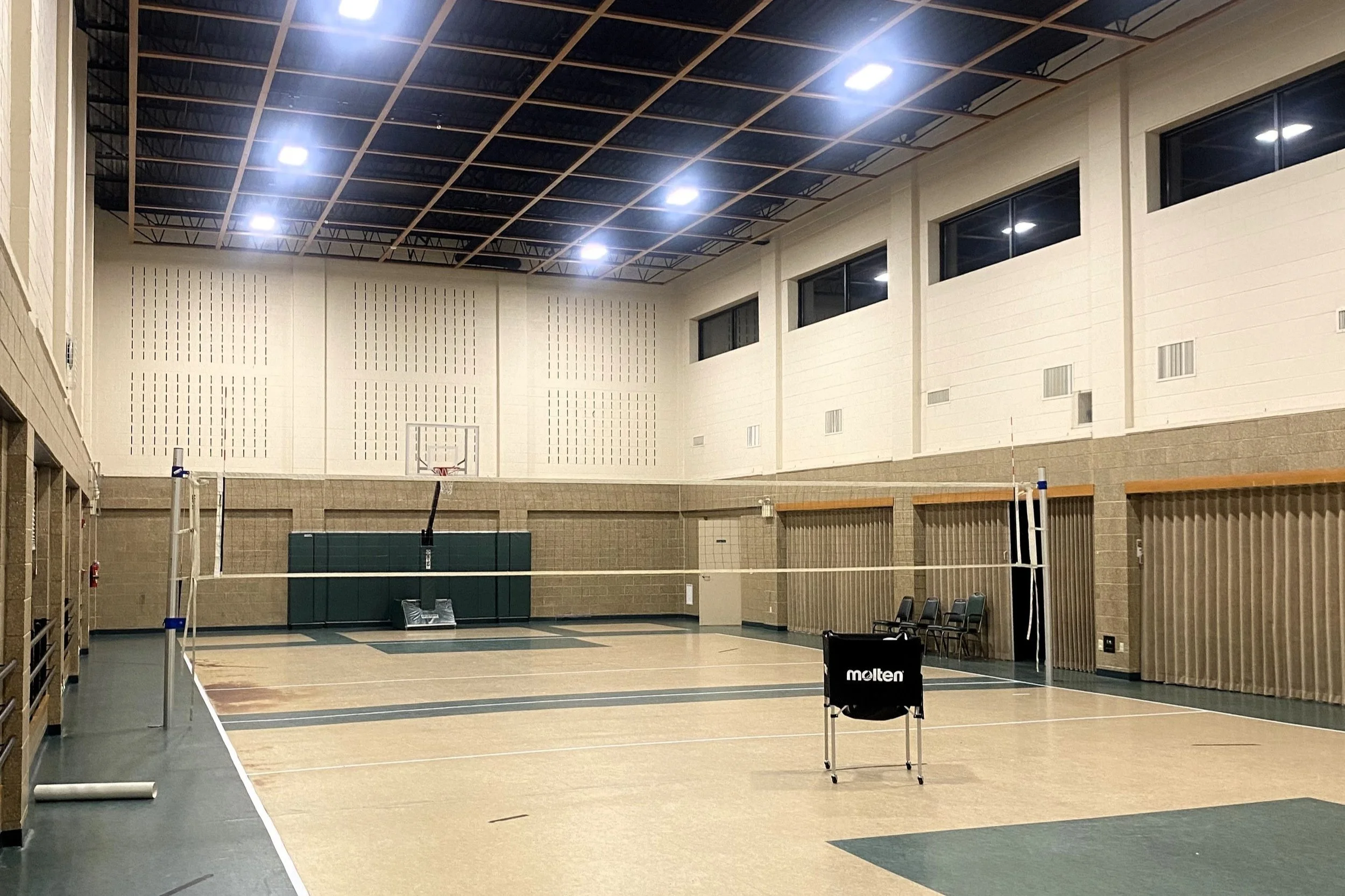 An indoor volleyball court with a net set up, a volleyball, a volleyball stand, chairs along the wall, and basketball hoop at the far end, inside a gym with high ceilings and bright lights.