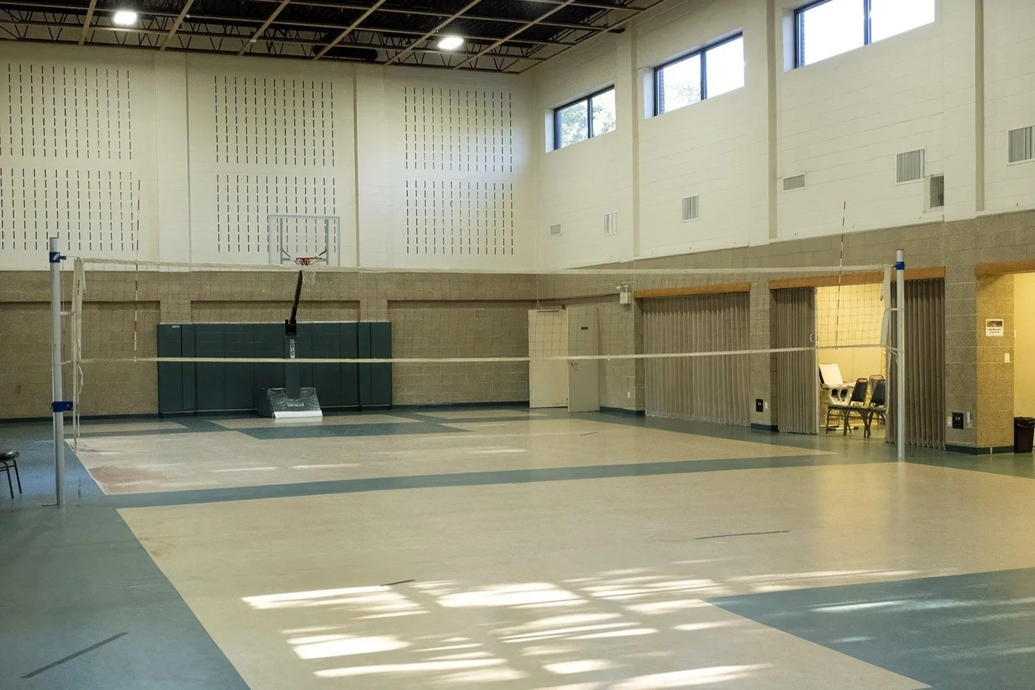 Empty indoor gymnasium with volleyball net, basketball hoop, and chairs along the wall.