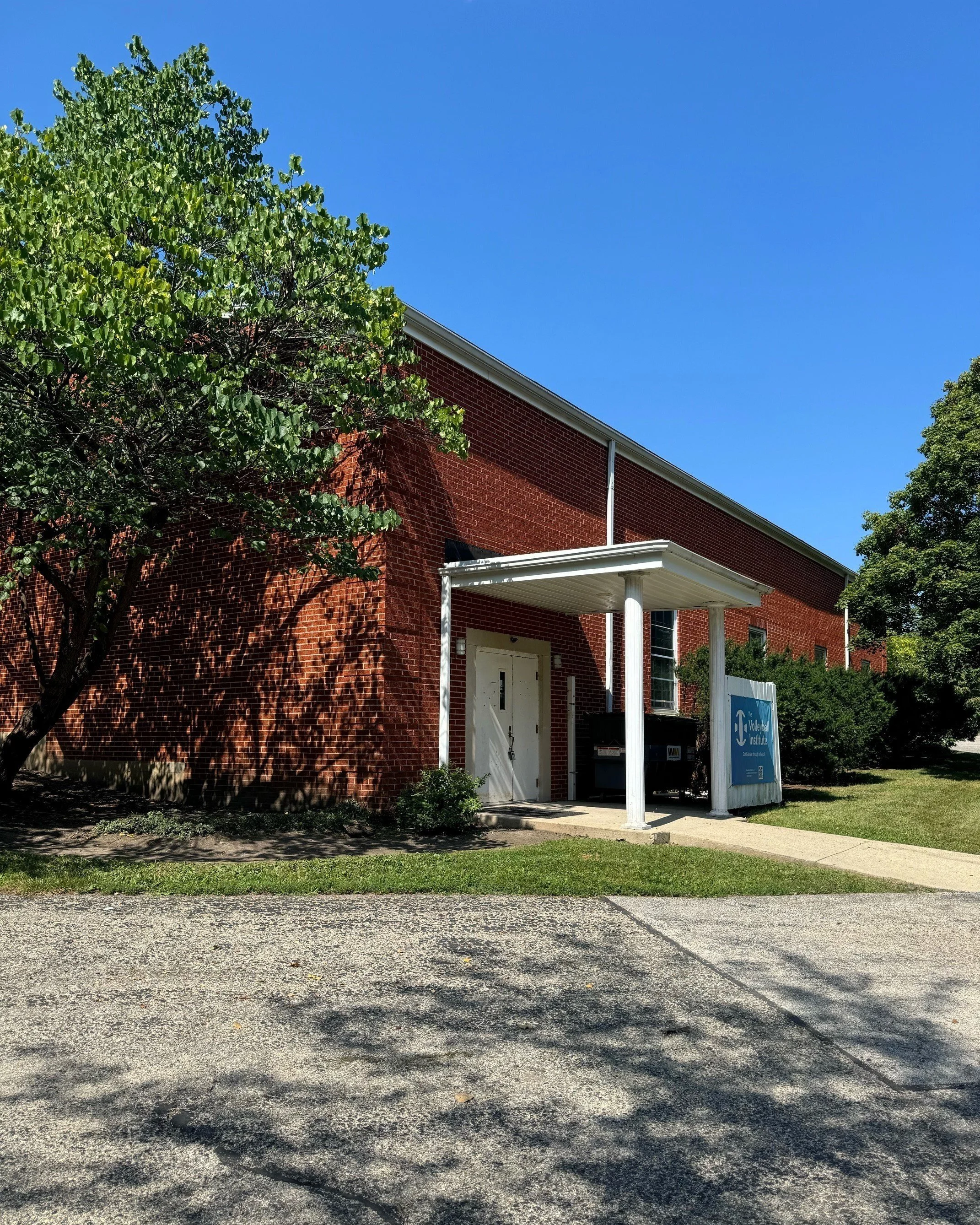 Red brick building with a white door, small porch, and a blue sign that reads 'Volunteer Intake.' A large leafy green tree and green bushes partially obscure the building, with a mix of sunny and shaded areas on the ground, and a clear blue sky overhead.
