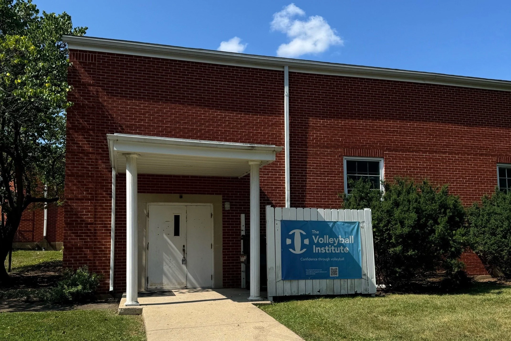 Brick building with a white door, a porch with white columns, and a sign for The Volleyball Institute outside.