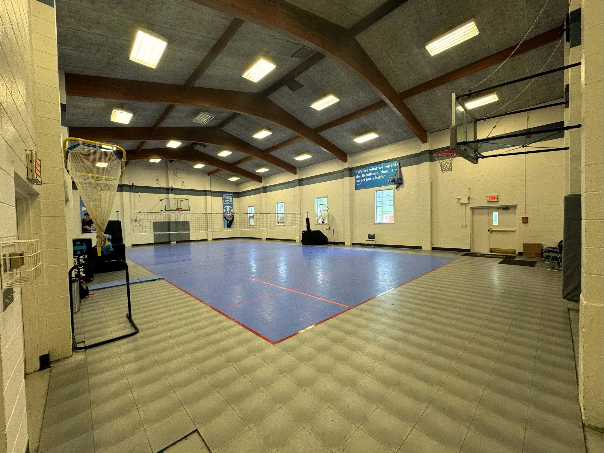 An indoor gymnasium with a blue volleyball court, basketball hoops, and a high ceiling with wooden beams, illuminated by fluorescent lights.