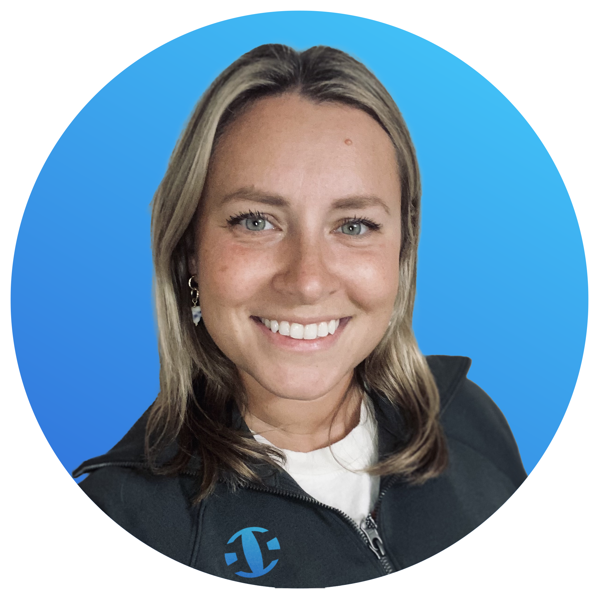 A smiling woman with blonde hair, wearing a black zip-up jacket with a logo on the chest, and a white shirt underneath, against a blue background.