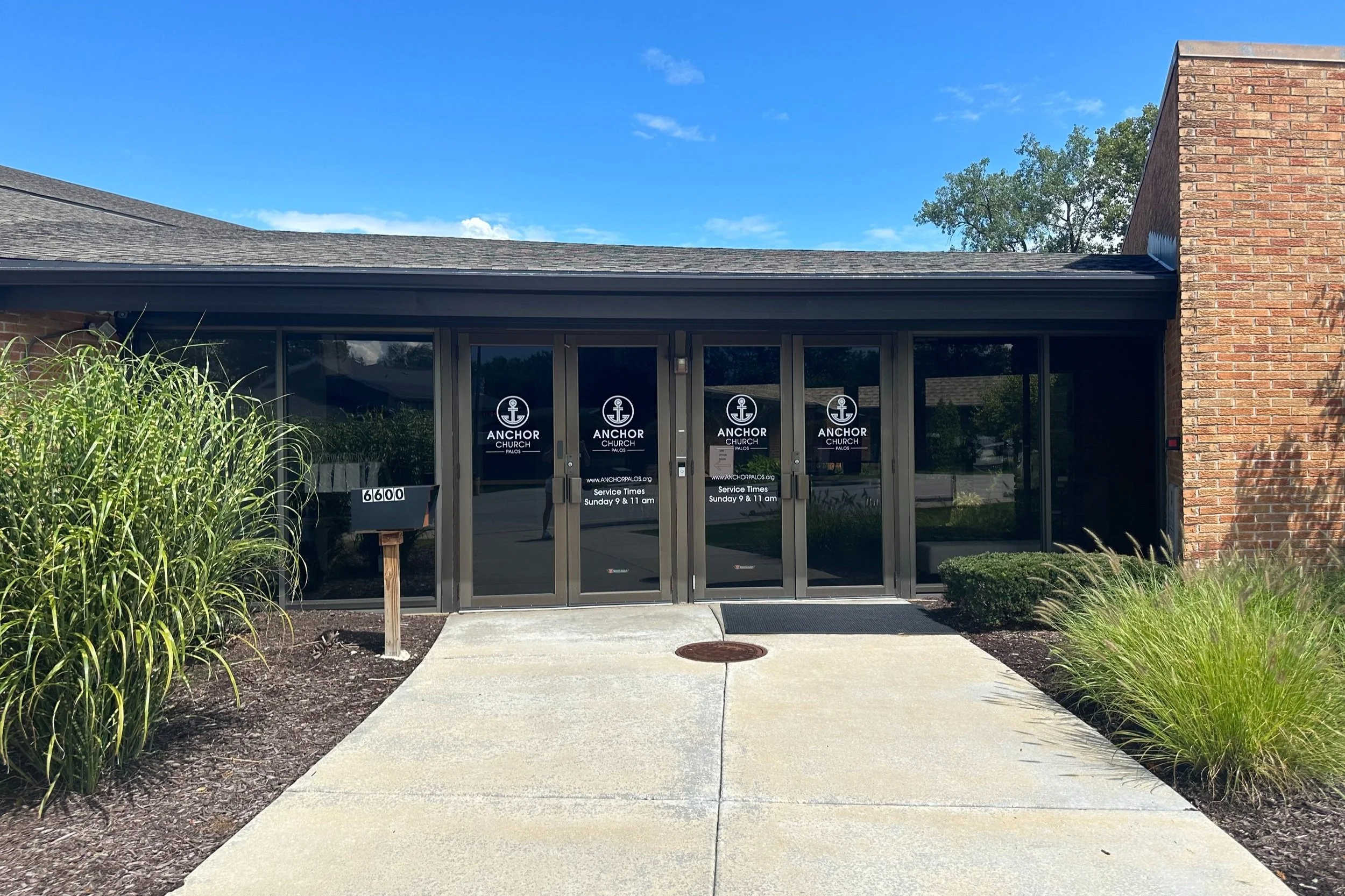 The entrance of a church building with glass doors displaying the church logo and service times, surrounded by landscaping with bushes and tall grass, and a sidewalk leading to the entrance.