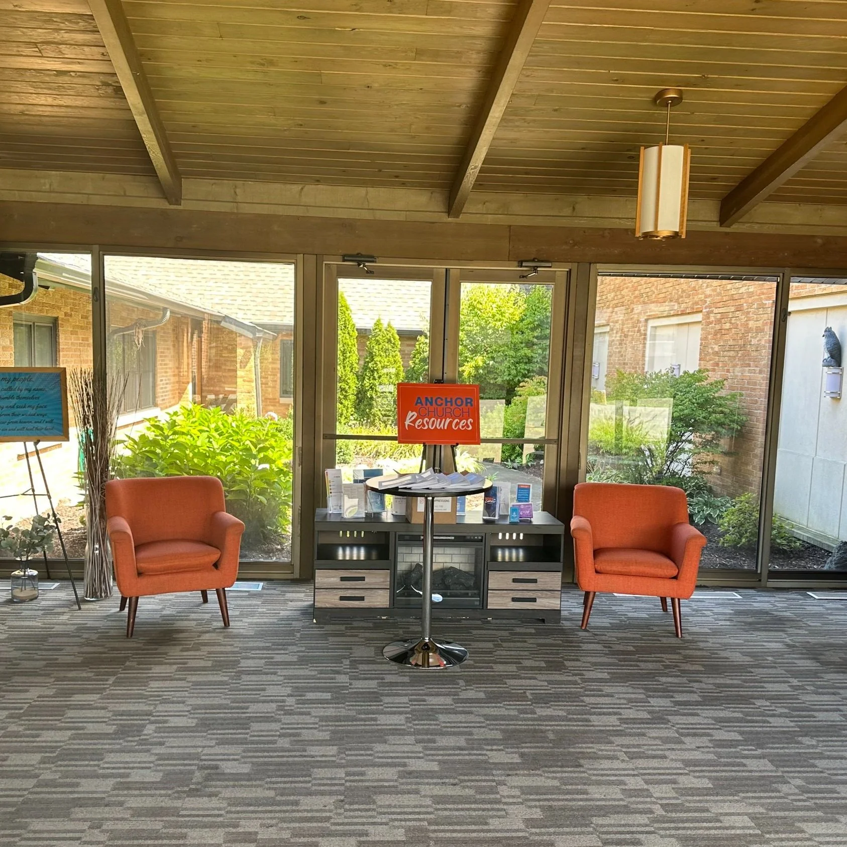 Indoor seating area with two orange armchairs, a small table, and a sign that reads 'Anchor Church Resources' on a stand, with large windows showing greenery outside.