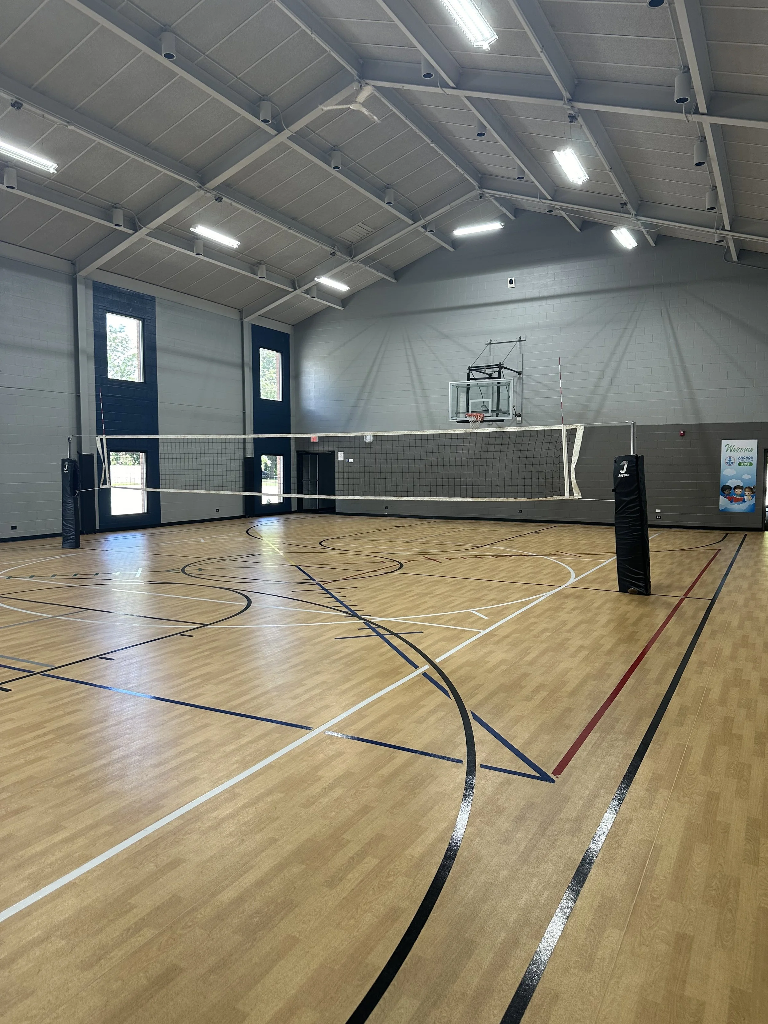 Indoor volleyball court with a volleyball net, basketball hoop in the background, and wooden flooring marked with various court lines.