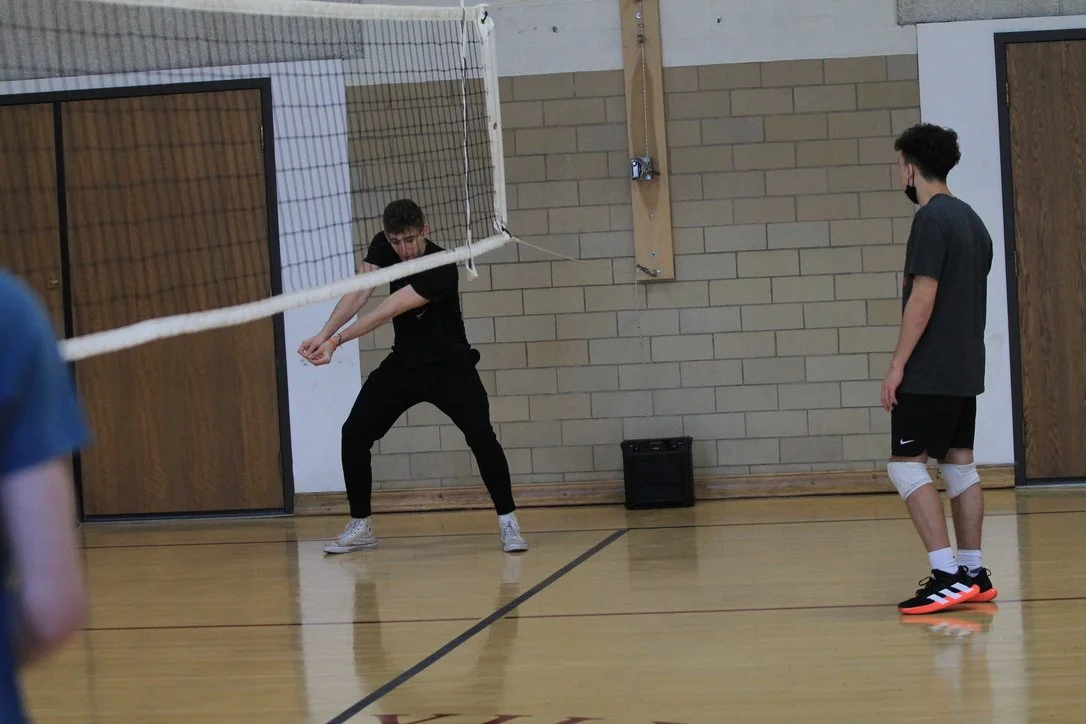 Two boys playing volleyball indoors, one is preparing to hit the ball while the other watches, all wearing casual athletic clothes and face masks.
