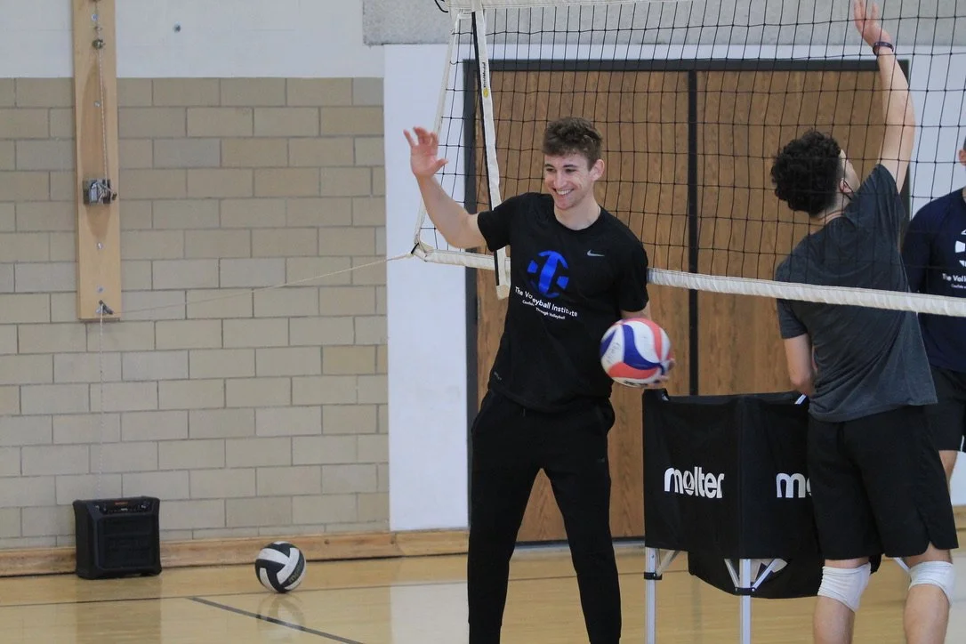 A young man in black sportswear holding a volleyball, smiling and waving, standing beside a volleyball net in a gymnasium with other players.