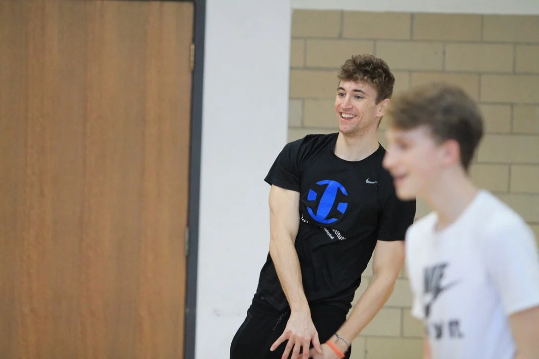 Two young men standing and laughing in a gym or classroom with brick and wood walls, wearing athletic T-shirts.
