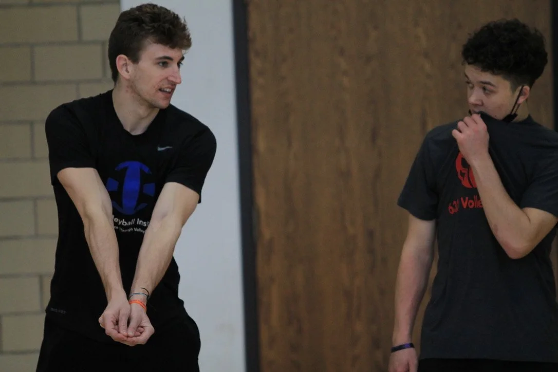 Two young men in sports attire having a conversation indoors, one with a volleyball logo on his shirt, the other with a mask pulled down from his face.