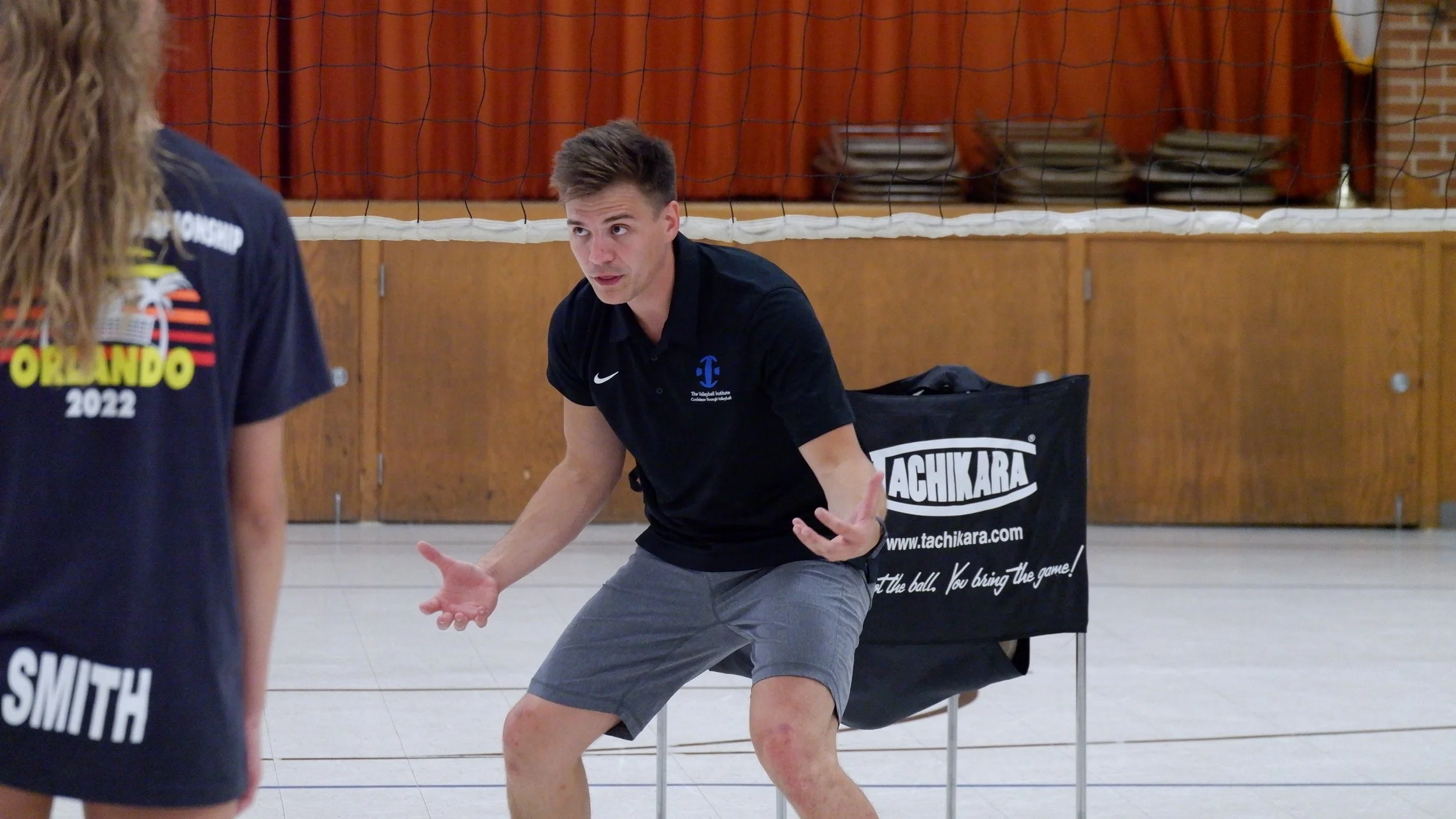 A male volleyball coach giving instructions during practice, with a volleyball net in the background.