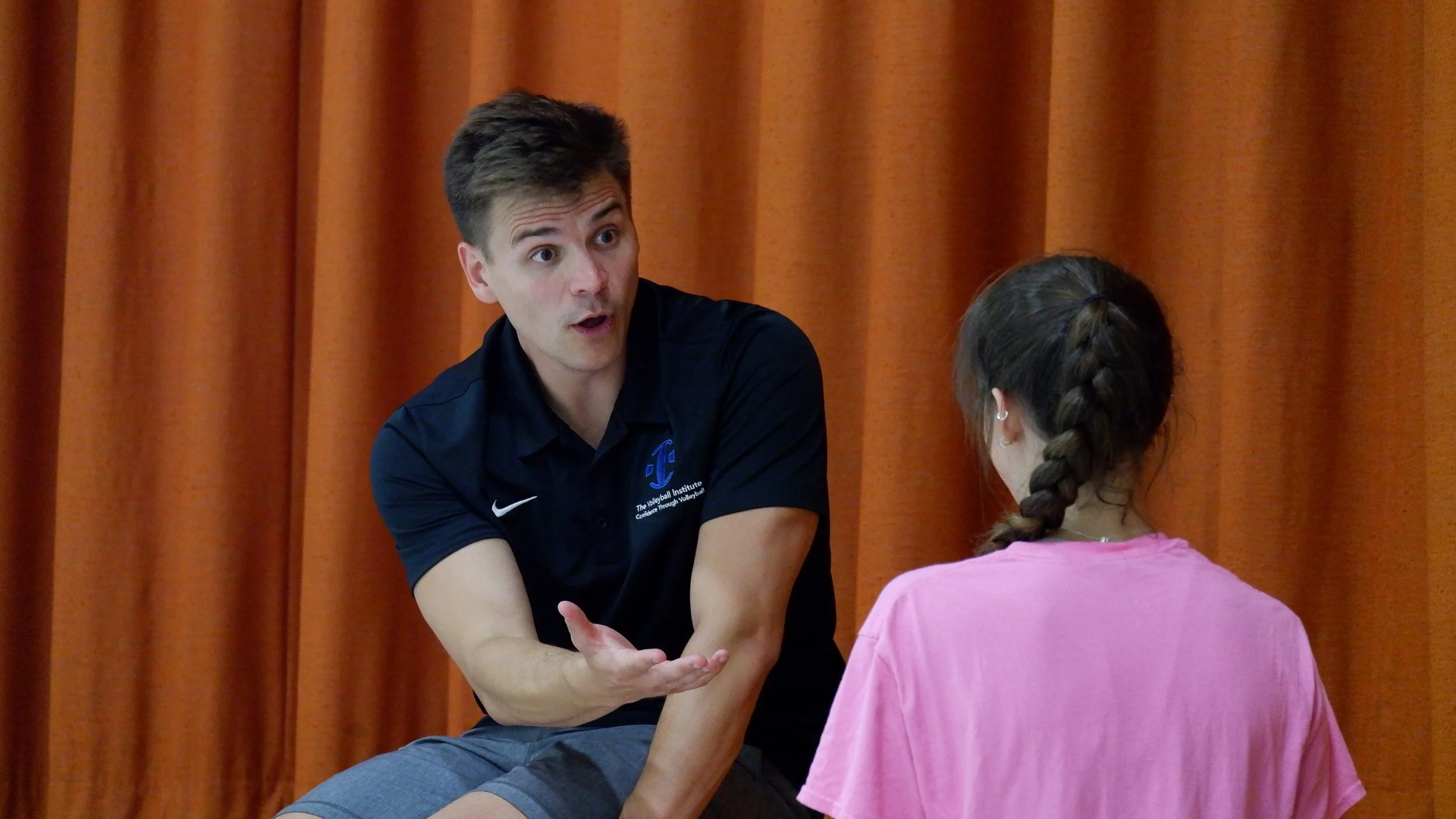 A man with short brown hair wearing a navy blue shirt engages in conversation with a young girl with braided hair wearing a pink shirt, seated in front of an orange curtain.