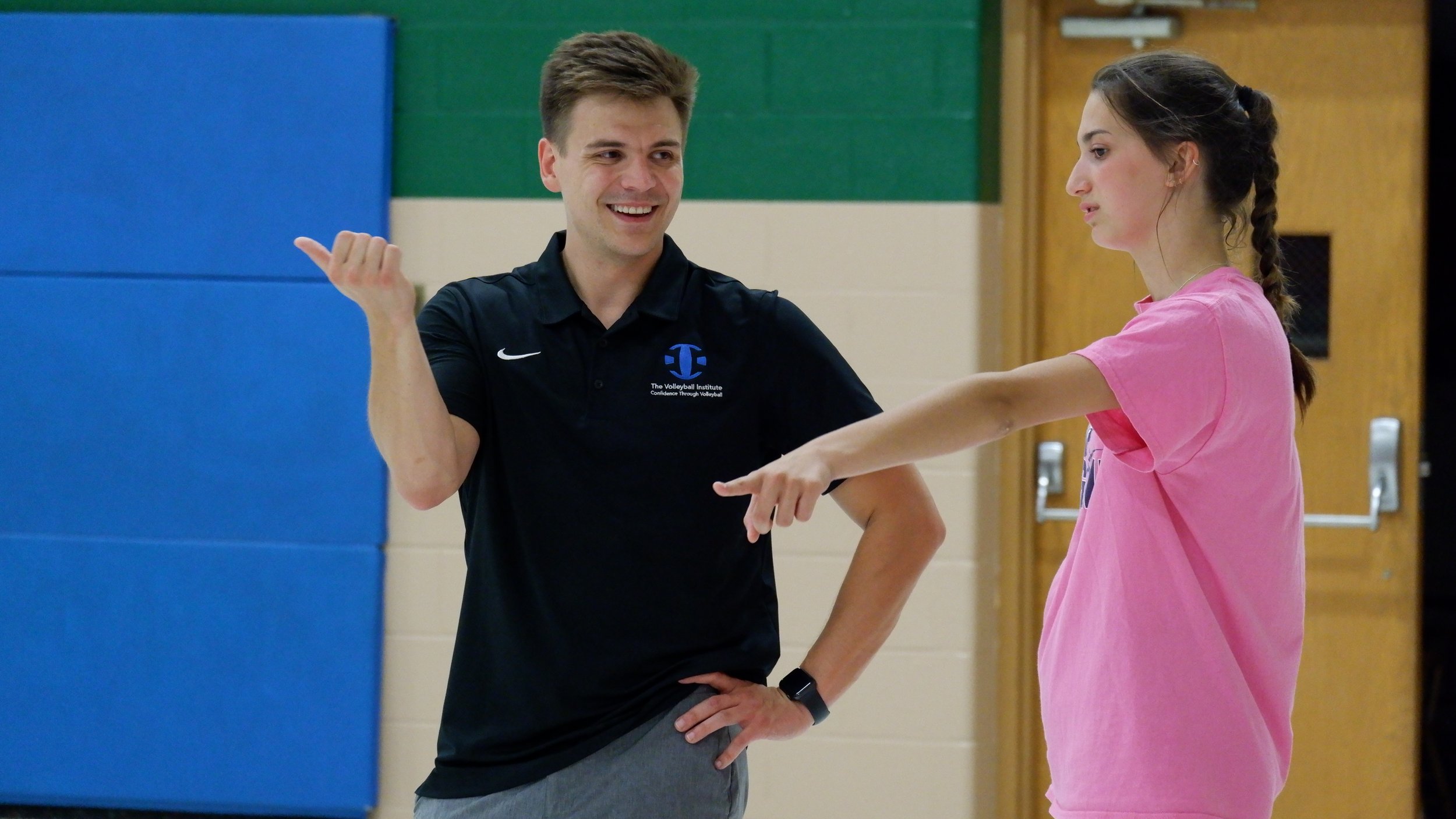 A male volleyball coach smiling and gesturing with his hand toward a young female player, who is focused on her arm and wearing a pink shirt, in a gymnasium.