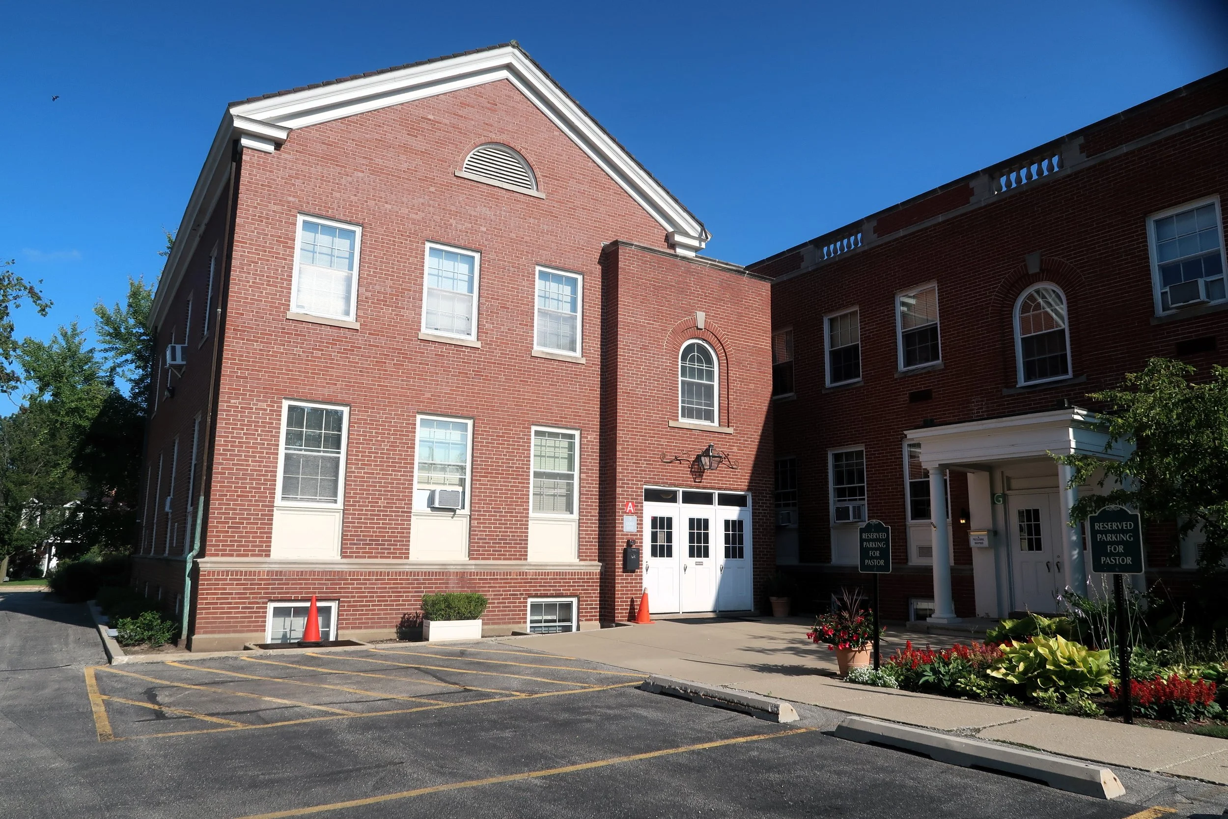 Red brick church building with white doors and windows, parking lot, and flowers in front, on a sunny day.